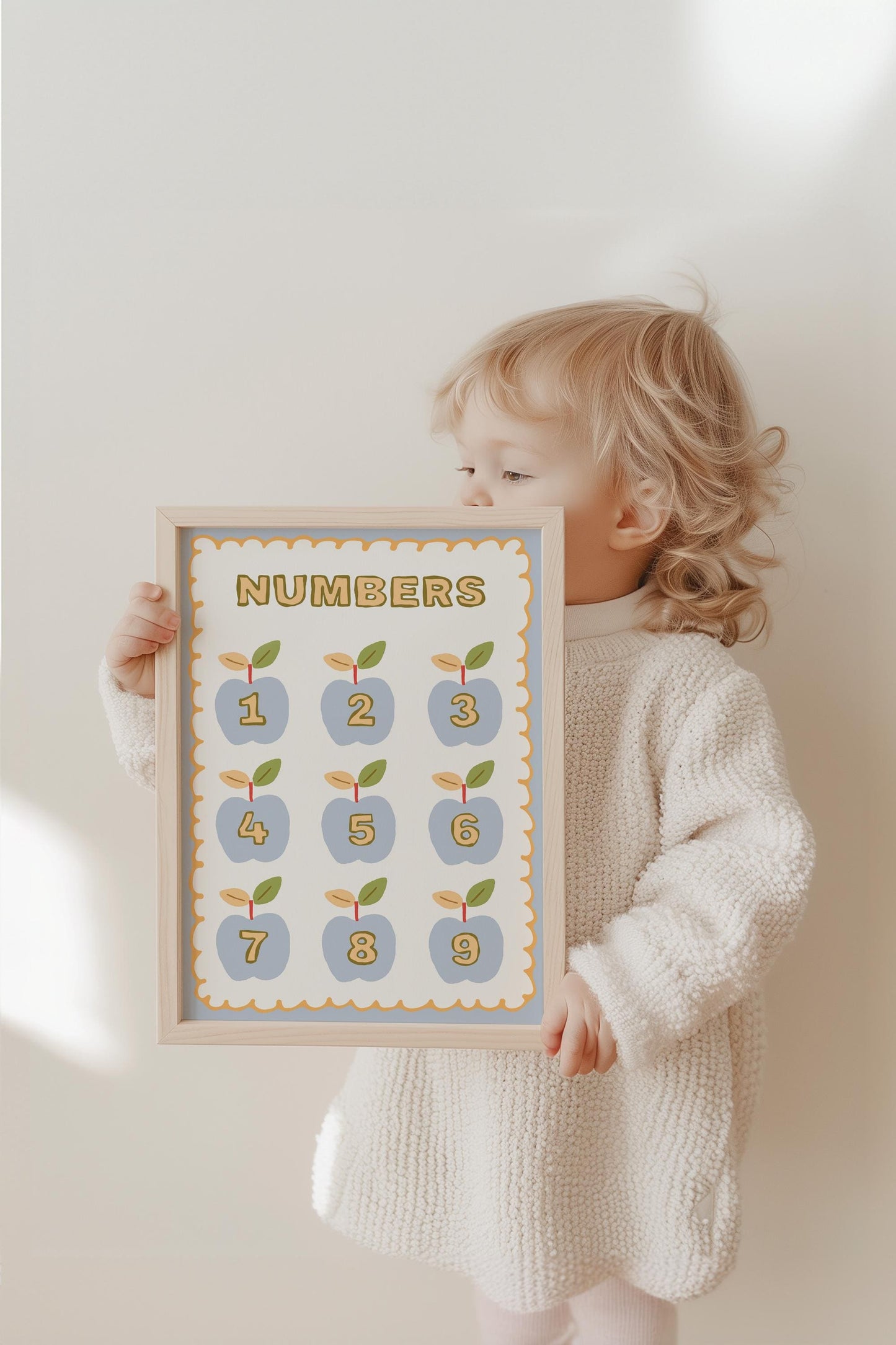 Ein kleines Kind hält das Poster Kinderzimmer Herzen von JUSTGOODMOOD, das Zahlen und Apfel-Design zeigt, perfekt für die frühkindliche Bildung.