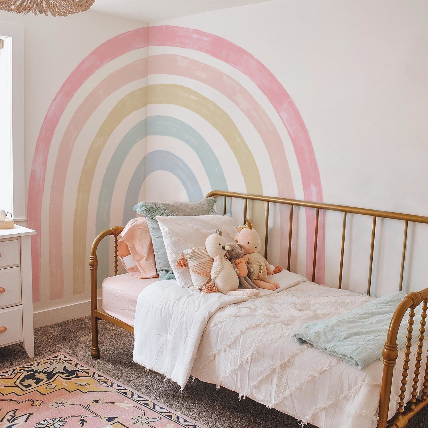 Niedliches Schlafzimmer mit Regenbogen-Wandaufkleber von justgoodmood. Ein farbenfrohes und einladendes Bett für Kinder, dekoriert mit Kuscheltieren und weichen Kissen.