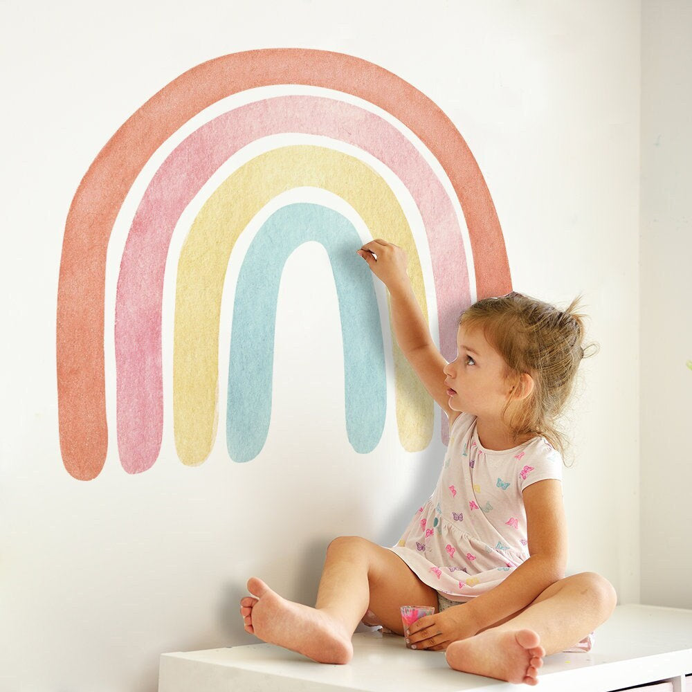 Ein kleines Mädchen dekoriert den Boho Stil Regenbogen Wandaufkleber von justgoodmood in ihrem Kinderzimmer. Perfekt für kreative Wandgestaltung und fröhliche Atmosphäre.