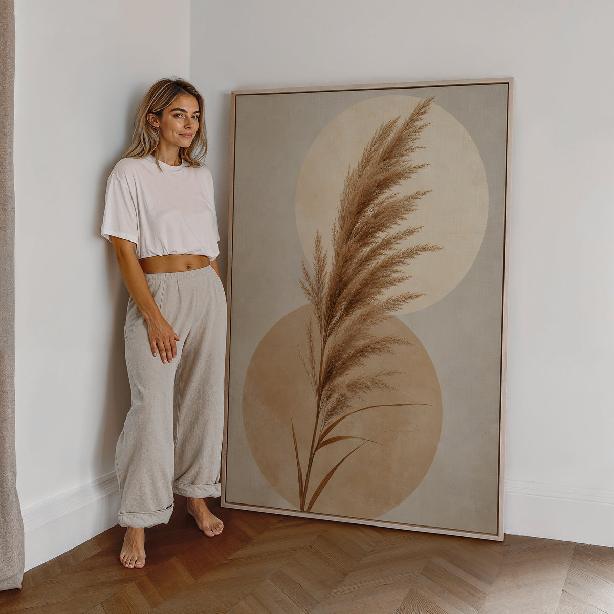 Motiv mit einem braunen Pampaswedel vor zwei überlappenden beigen Kreisen, als großes gerahmtes Wandbild neben einer Frau.