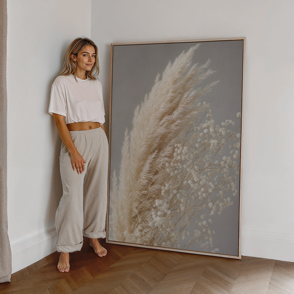 Motiv mit cremeweißen Pampaswedeln neben zartem weißen Schleierkraut vor grauem Hintergrund, als großes gerahmtes Wandbild neben einer Frau.