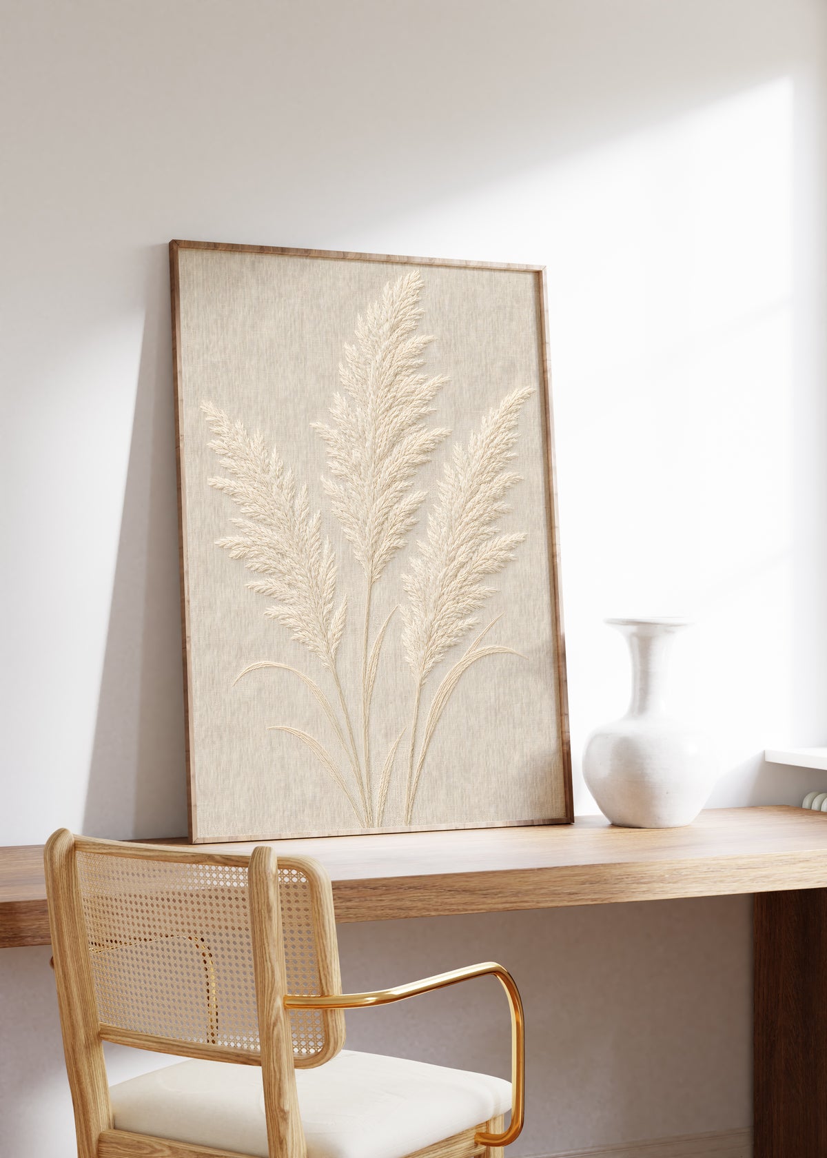 Gerahmtes Poster mit drei cremeweißen Pampaswedeln in Reliefoptik auf beigefarbenem Hintergrund, auf einem Holztisch neben einer weißen Vase.