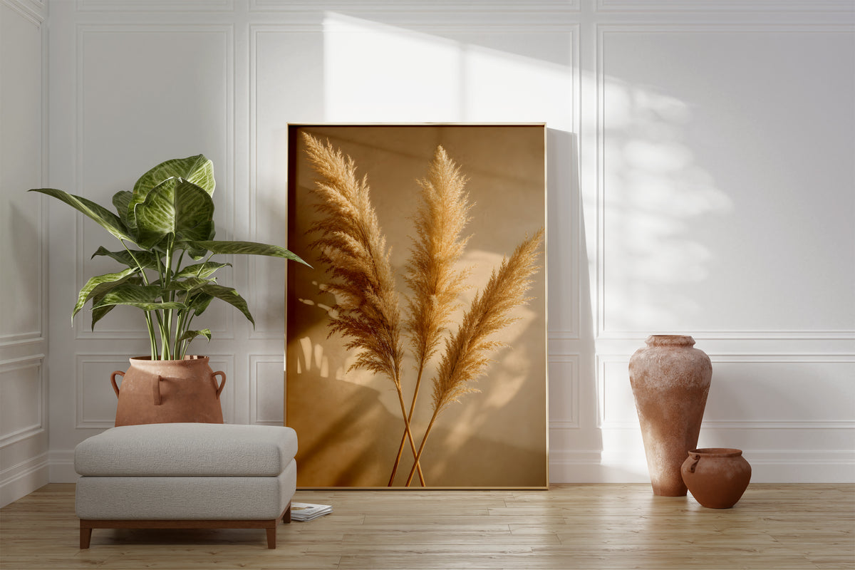 Motiv mit drei goldenen Pampaswedeln mit langen Schatten auf warmbeigem Hintergrund, als gerahmtes Poster neben Sessel und Zimmerpflanze.