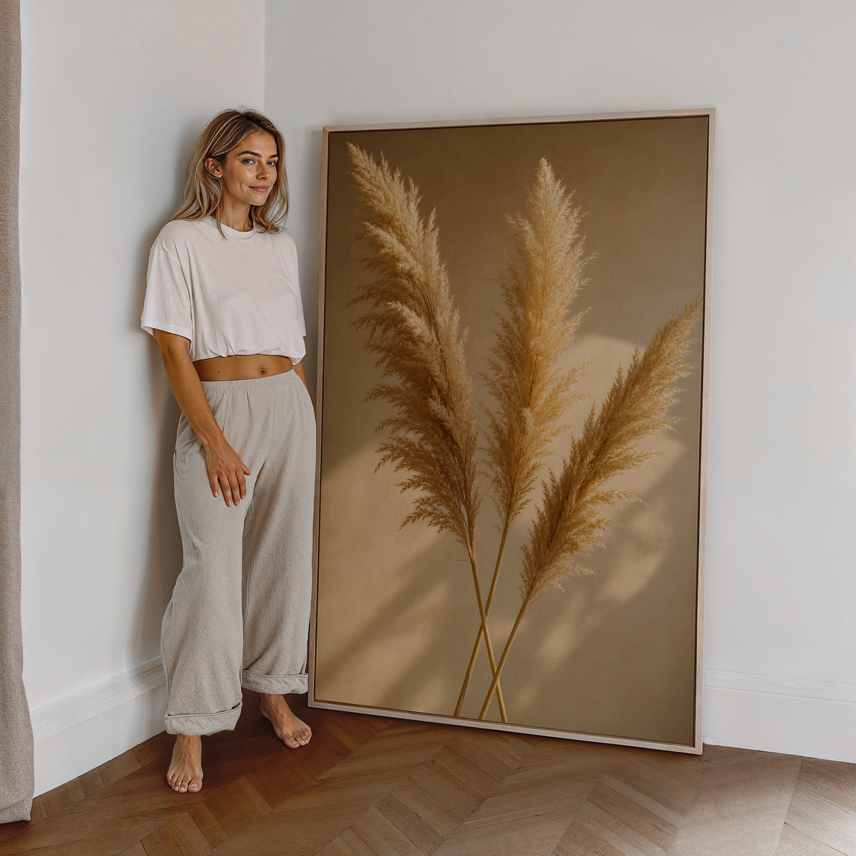 Motiv mit drei goldenen Pampaswedeln mit langen Schatten auf warmbeigem Hintergrund, als großes gerahmtes Wandbild neben einer Frau.