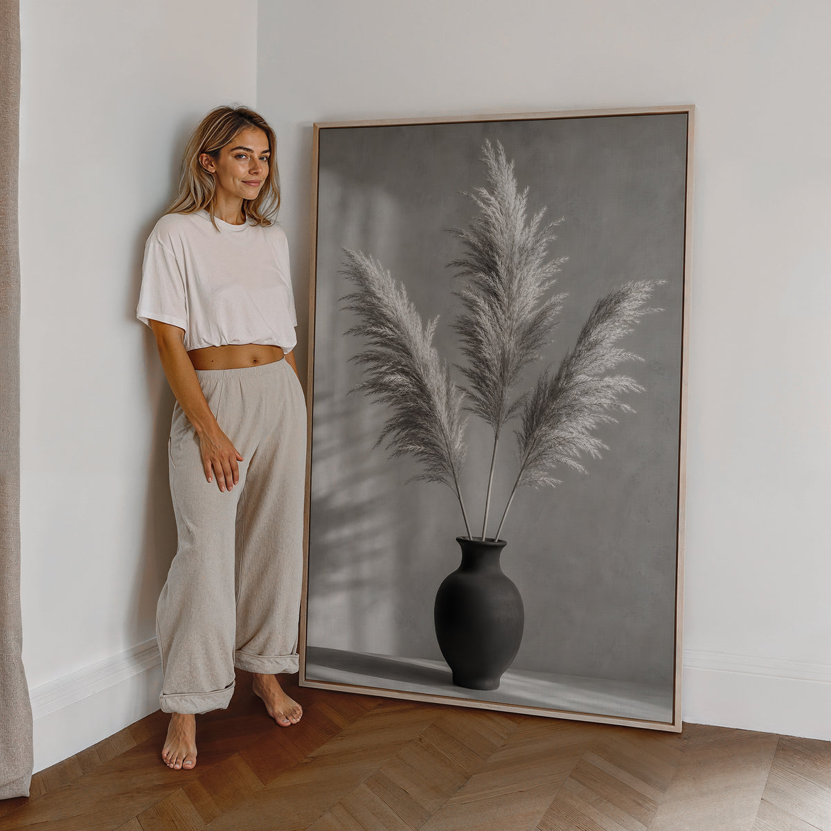 Motiv mit drei Pampaswedeln in einer schwarzen Vase vor einer grauen Wand mit Schatten, als großes gerahmtes Wandbild neben einer Frau.