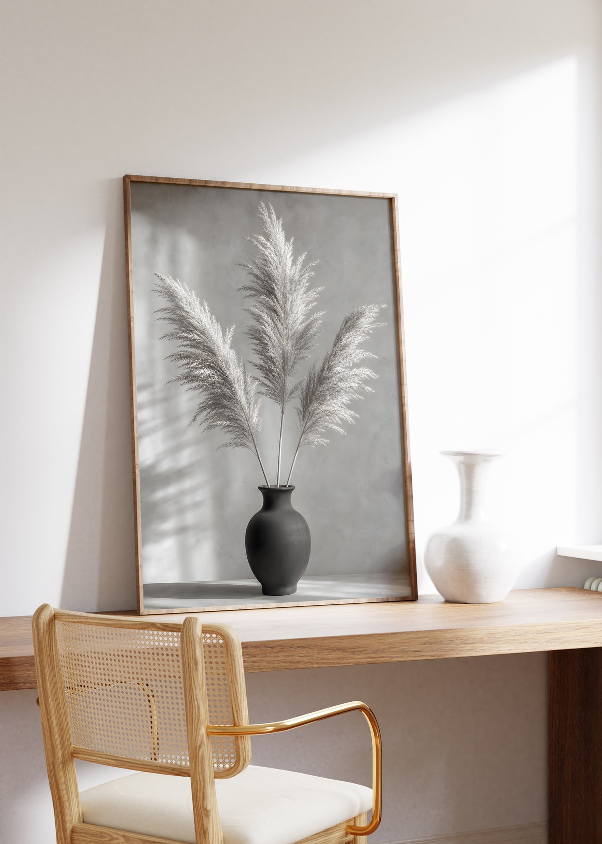 Gerahmtes Poster mit drei Pampaswedeln in einer schwarzen Vase vor einer grauen Wand mit Schatten, auf einem Holztisch neben einer weißen Vase.