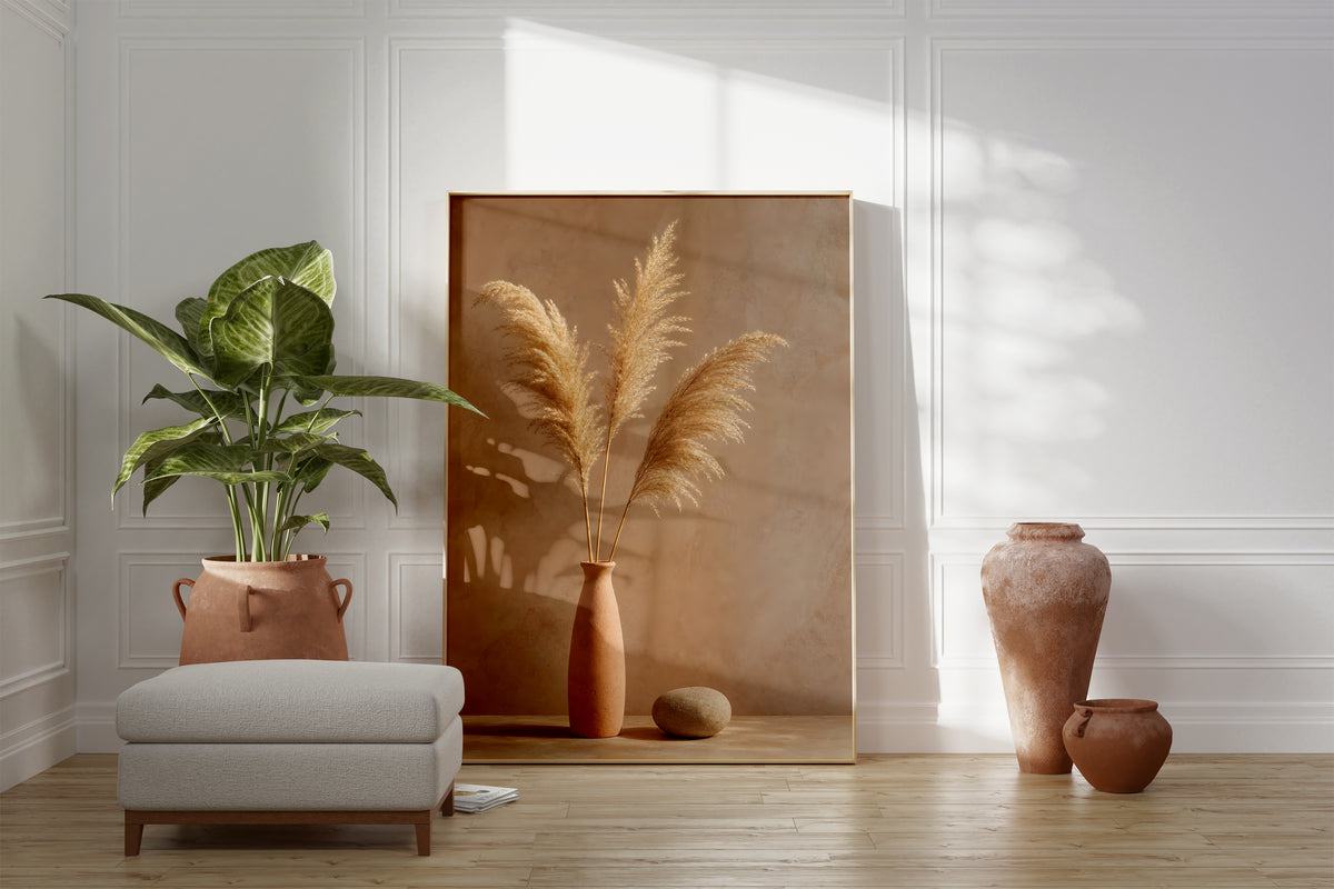 Motiv mit drei goldbraunen Pampaswedeln in einer schmalen Terrakottavase neben einem Stein, als gerahmtes Poster neben Sessel und Zimmerpflanze.