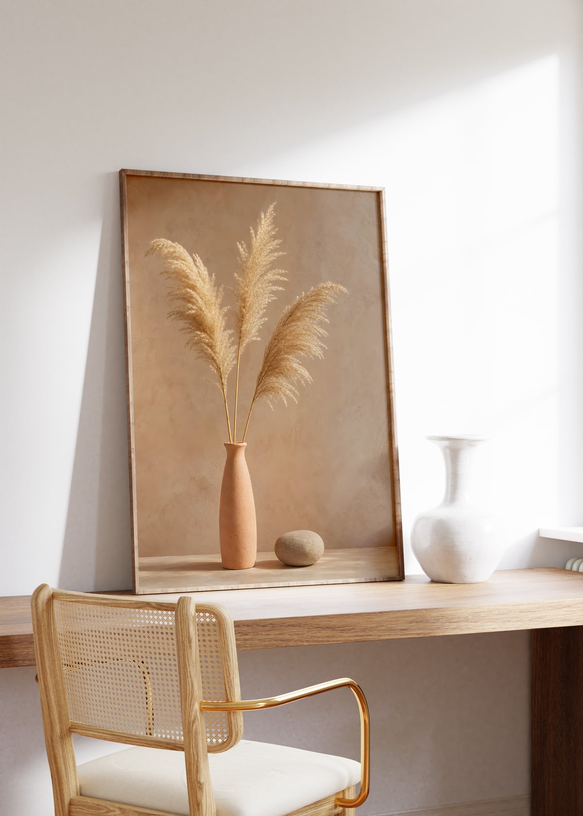 Gerahmtes Poster mit drei goldbraunen Pampaswedeln in einer schmalen Terrakottavase neben einem Stein, auf einem Holztisch neben einer weißen Vase.
