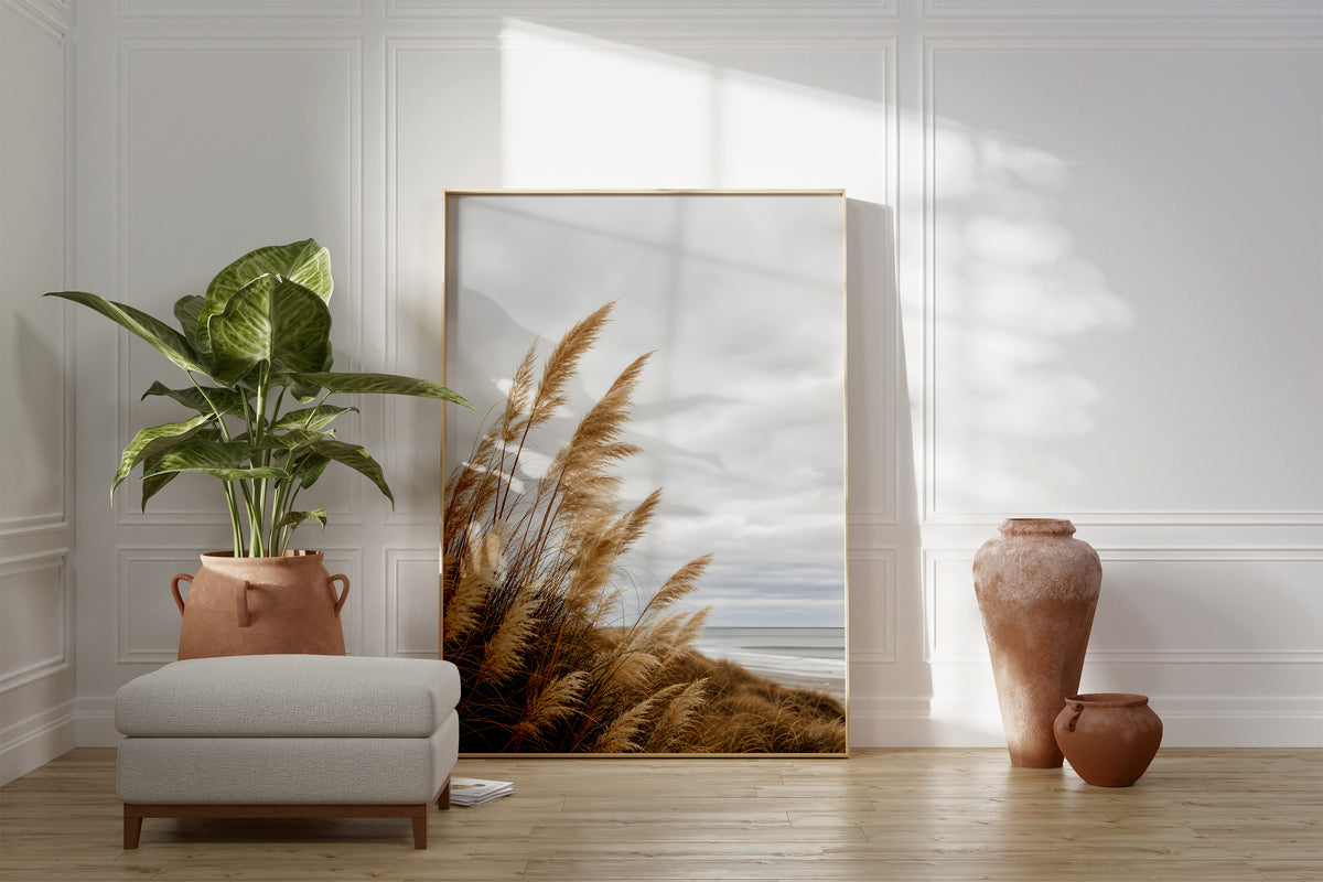 Motiv mit goldbraunem Dünengras am Strand unter grauen Wolken, als gerahmtes Poster neben Sessel und Zimmerpflanze.