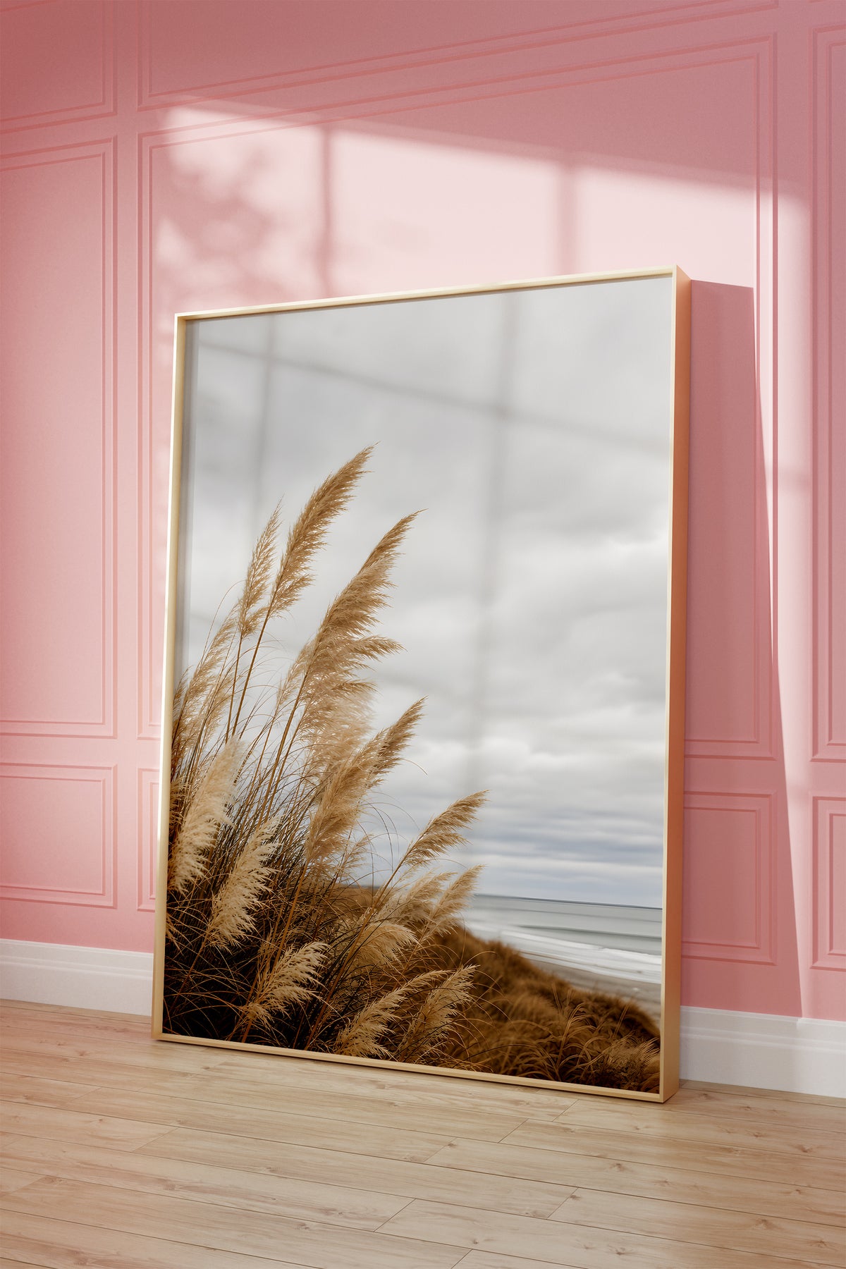 Motiv mit goldbraunem Dünengras am Strand unter grauen Wolken, als gerahmtes Poster vor einer rosafarbenen Wand.