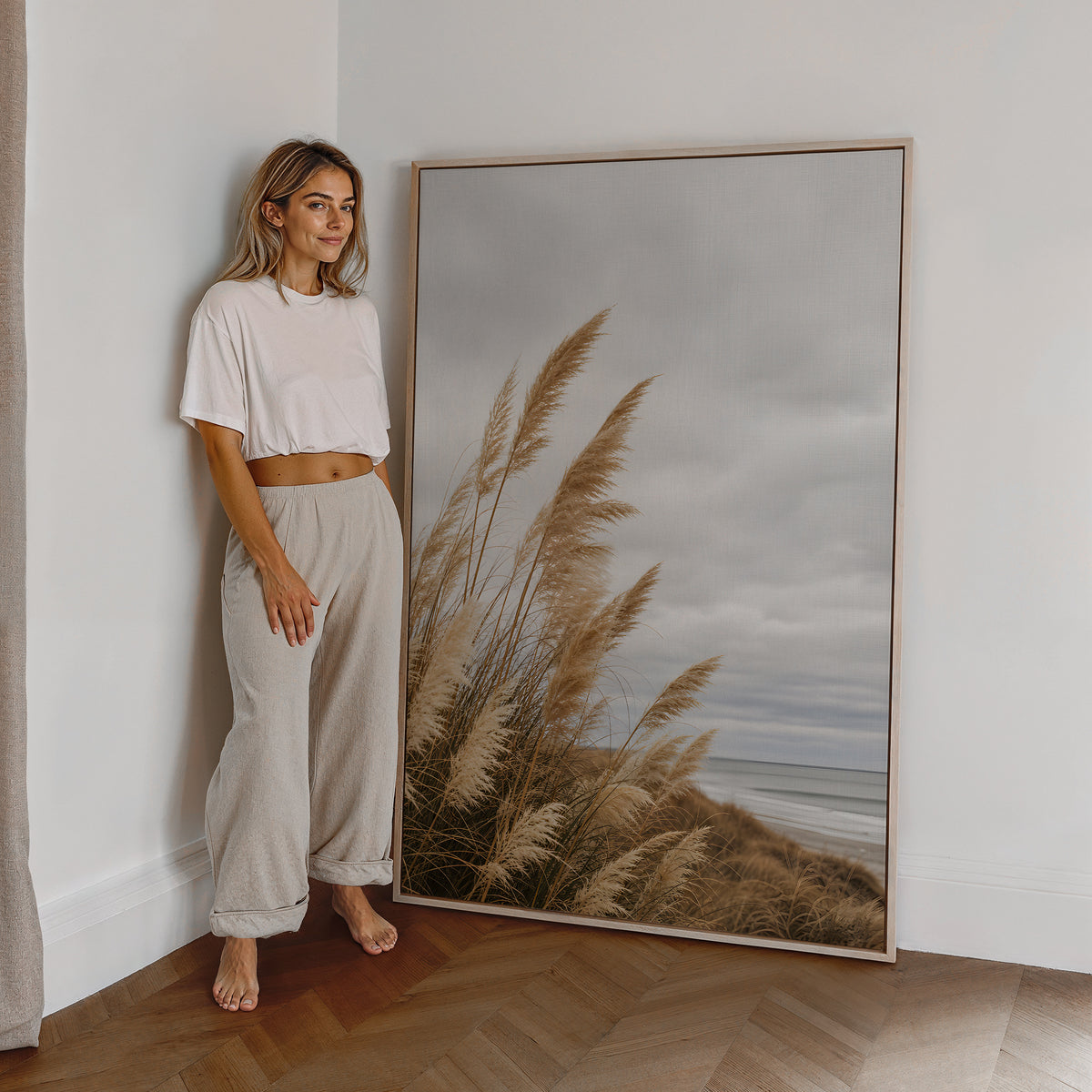 Motiv mit goldbraunem Dünengras am Strand unter grauen Wolken, als großes gerahmtes Wandbild neben einer Frau.