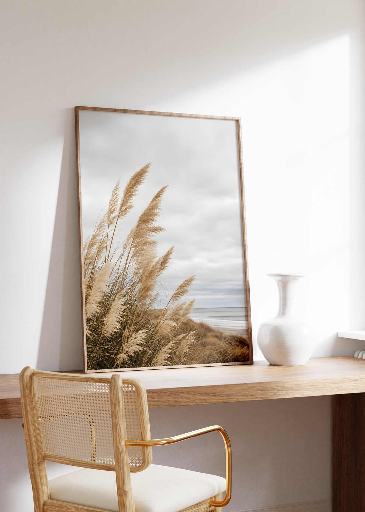 Gerahmtes Poster mit goldbraunem Dünengras am Strand unter grauen Wolken, auf einem Holztisch neben einer weißen Vase.