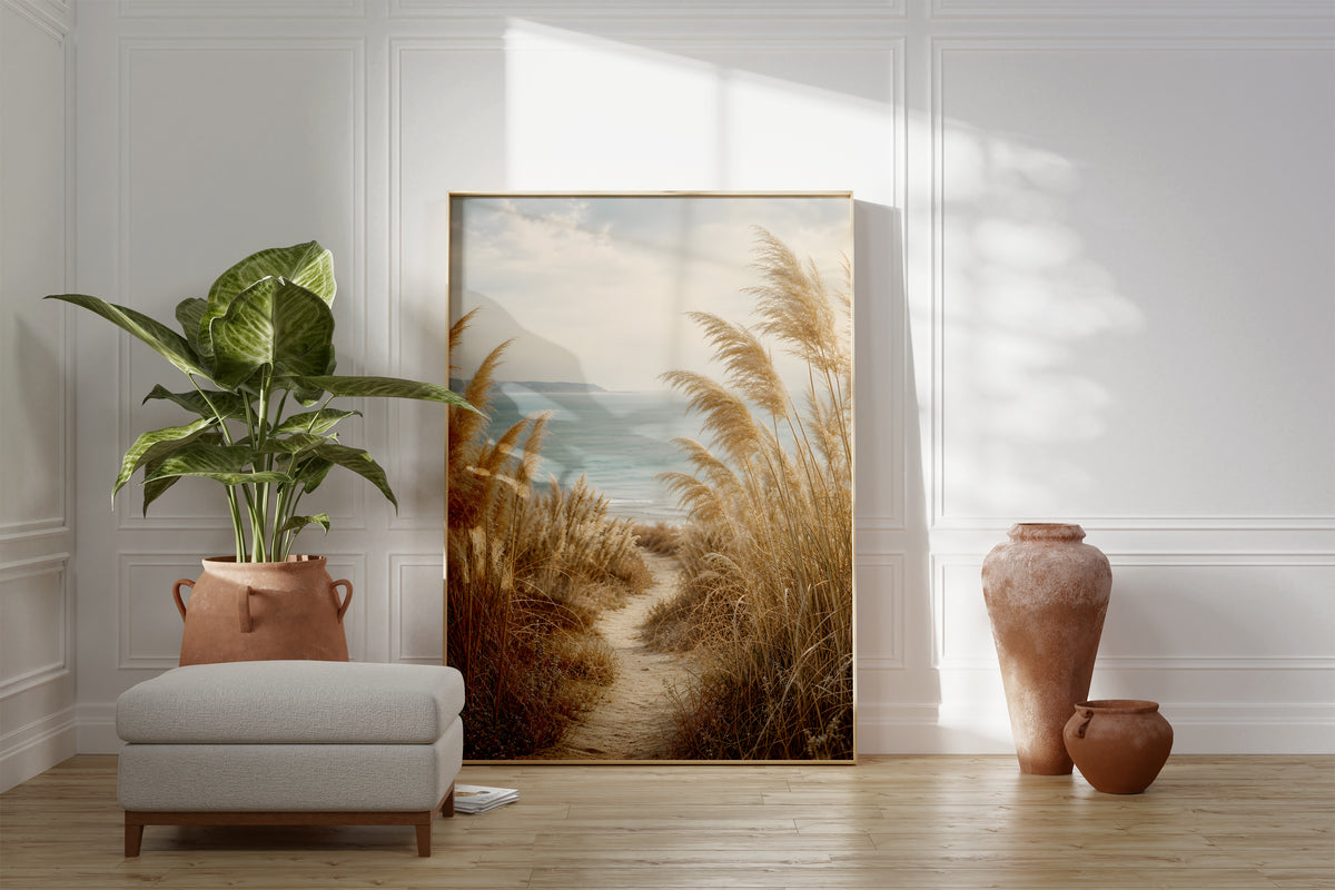 Motiv mit einem schmalen Sandpfad zwischen Pampasgräsern zum blauen Meer, als gerahmtes Poster neben Sessel und Zimmerpflanze.