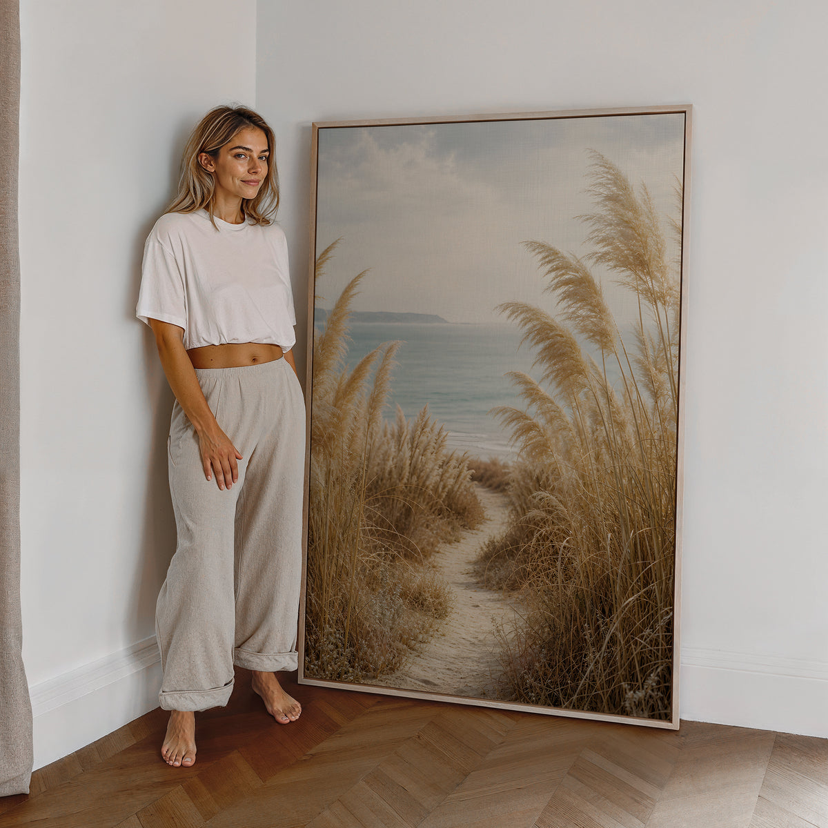 Motiv mit einem schmalen Sandpfad zwischen Pampasgräsern zum blauen Meer, als großes gerahmtes Wandbild neben einer Frau.