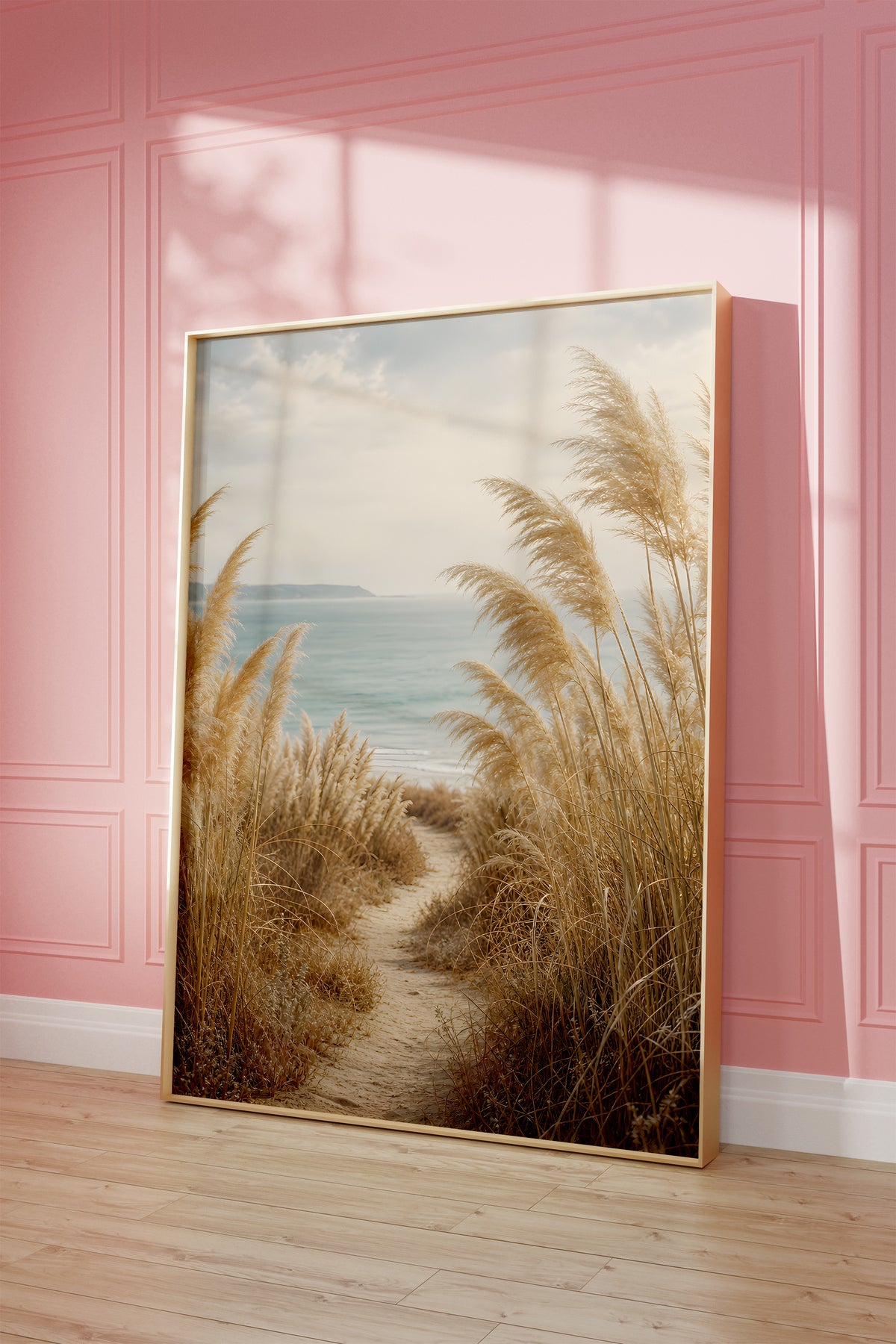 Motiv mit einem schmalen Sandpfad zwischen Pampasgräsern zum blauen Meer, als gerahmtes Poster vor einer rosafarbenen Wand.