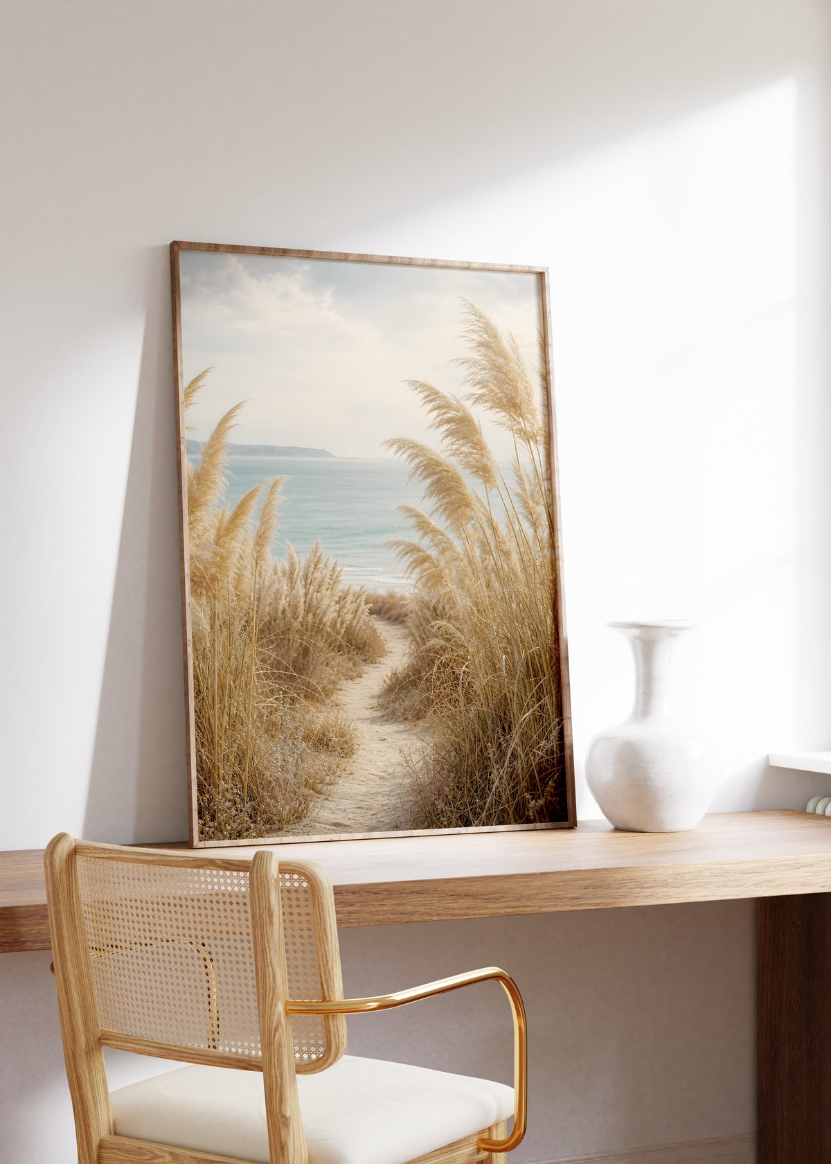 Gerahmtes Poster mit einem schmalen Sandpfad zwischen Pampasgräsern zum blauen Meer, auf einem Holztisch neben einer weißen Vase.