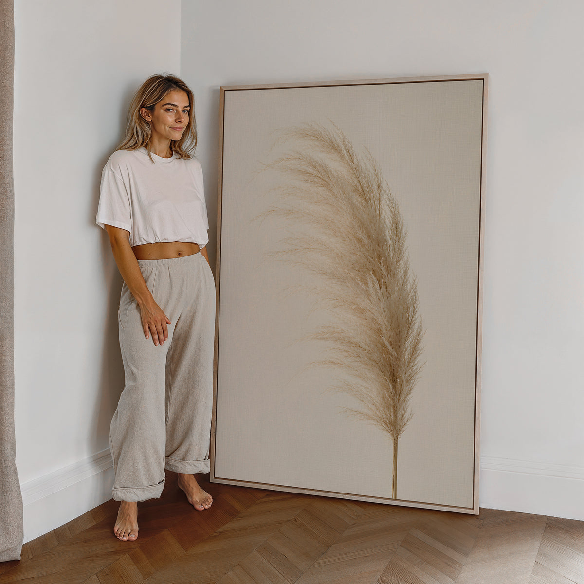 Motiv mit einem einzelnen flauschigen Pampaswedel in Cremebeige vor hellem Hintergrund, als großes gerahmtes Wandbild neben einer Frau.
