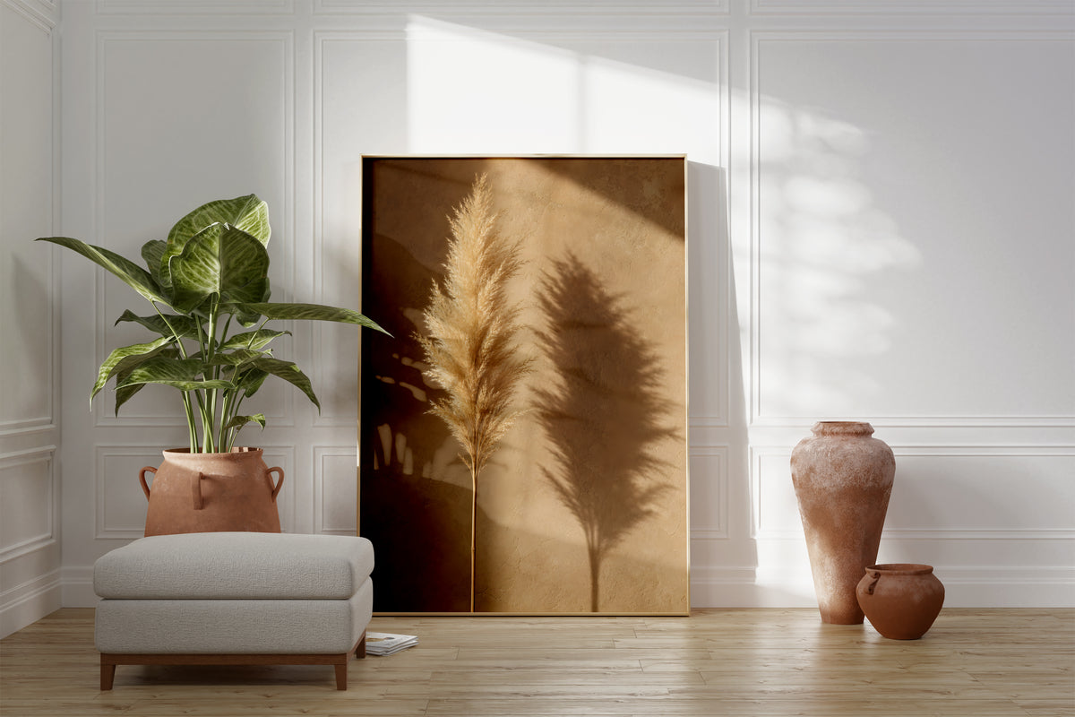 Motiv mit einem goldbeigen Pampaswedel und seinem Schatten auf einer warmbraunen Wand, als gerahmtes Poster neben Sessel und Zimmerpflanze.