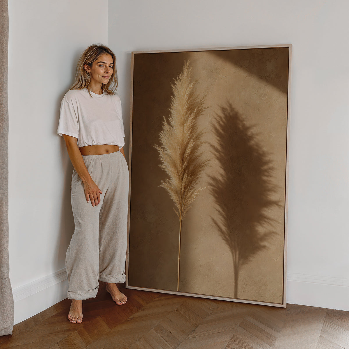 Motiv mit einem goldbeigen Pampaswedel und seinem Schatten auf einer warmbraunen Wand, als großes gerahmtes Wandbild neben einer Frau.