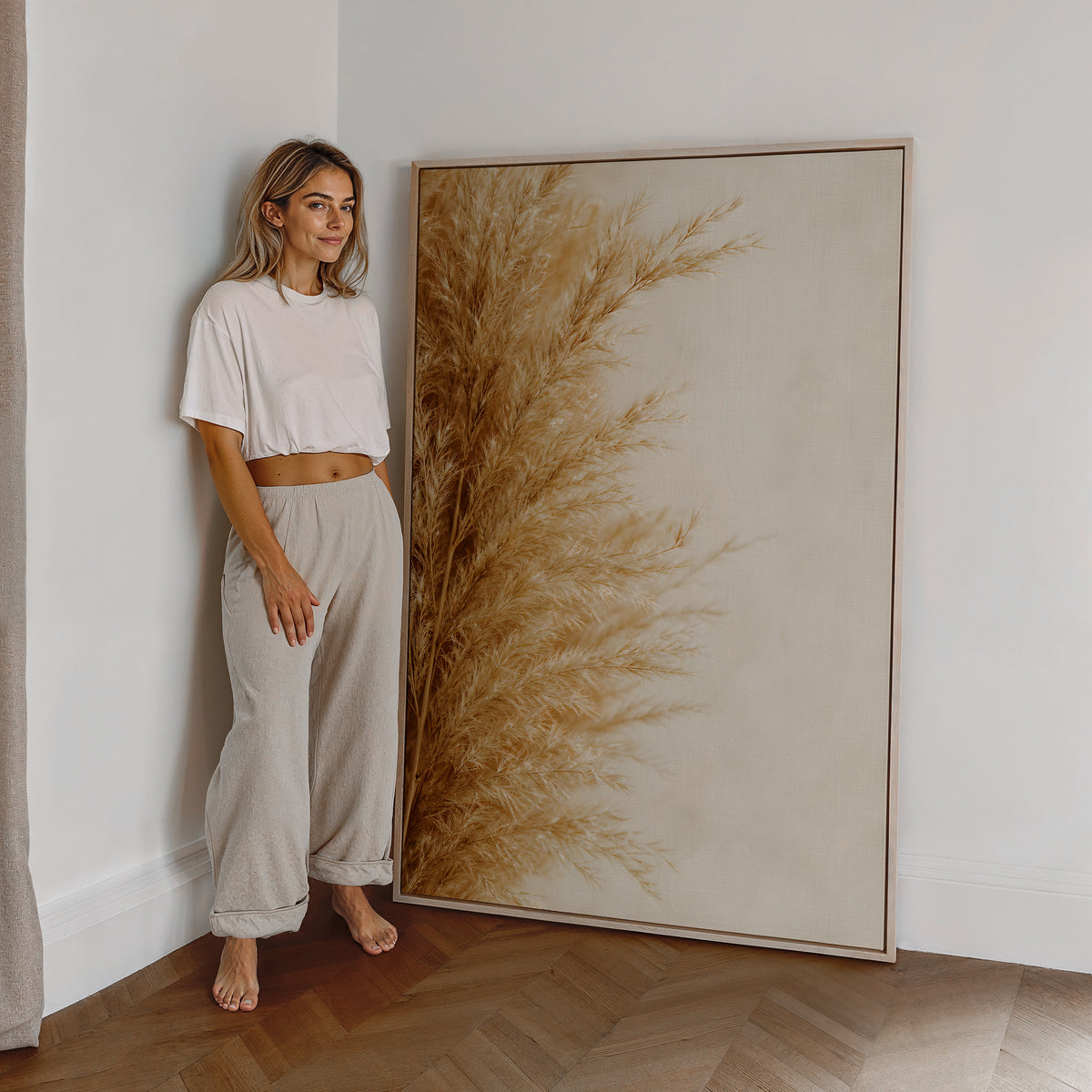 Motiv mit goldbeigen Pampasfasern am linken Rand vor hellem Hintergrund, als großes gerahmtes Wandbild neben einer Frau.