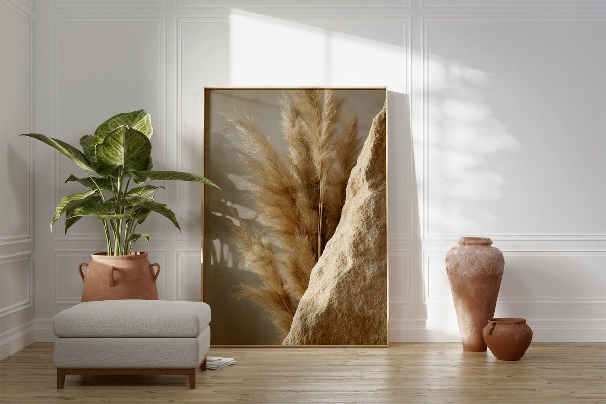 Motiv mit goldbraunen Pampaswedeln neben einer rauen sandfarbenen Steinfläche, als gerahmtes Poster neben Sessel und Zimmerpflanze.