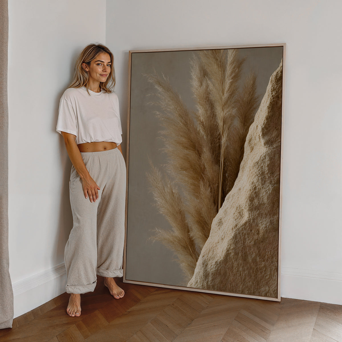Motiv mit goldbraunen Pampaswedeln neben einer rauen sandfarbenen Steinfläche, als großes gerahmtes Wandbild neben einer Frau.