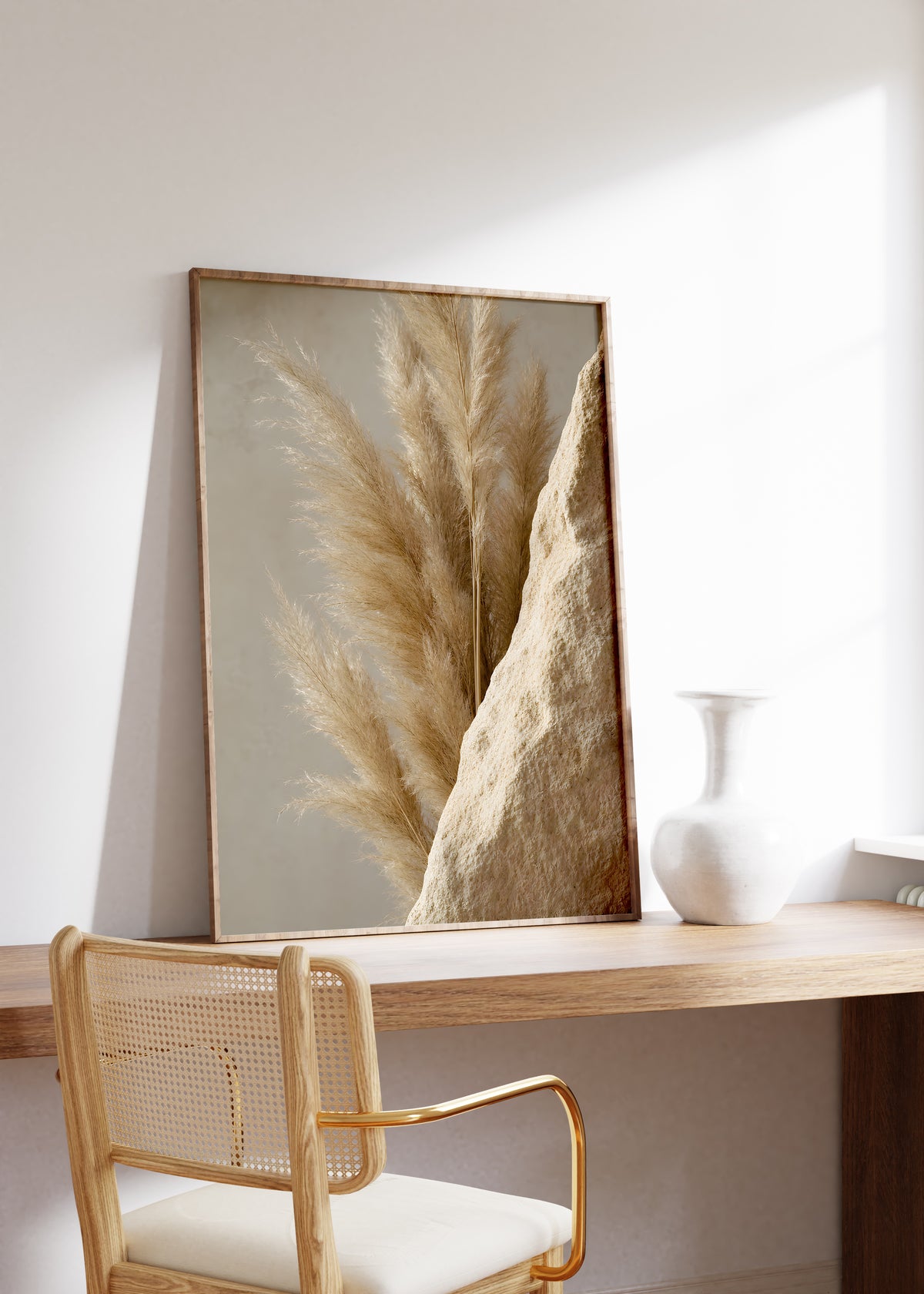 Gerahmtes Poster mit goldbraunen Pampaswedeln neben einer rauen sandfarbenen Steinfläche, auf einem Holztisch neben einer weißen Vase.