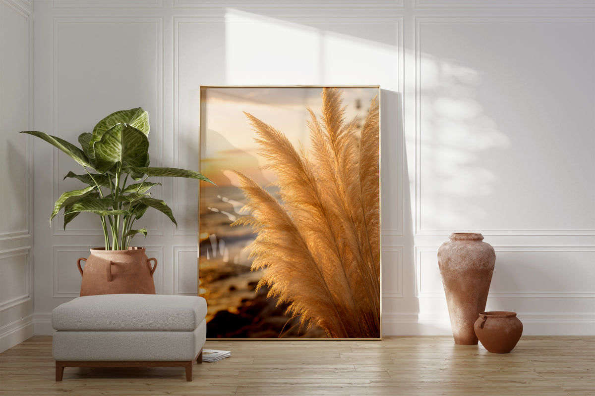 Motiv mit goldenen Pampaswedeln in Nahaufnahme am Strand im warmen Abendlicht, als gerahmtes Poster neben Sessel und Zimmerpflanze.