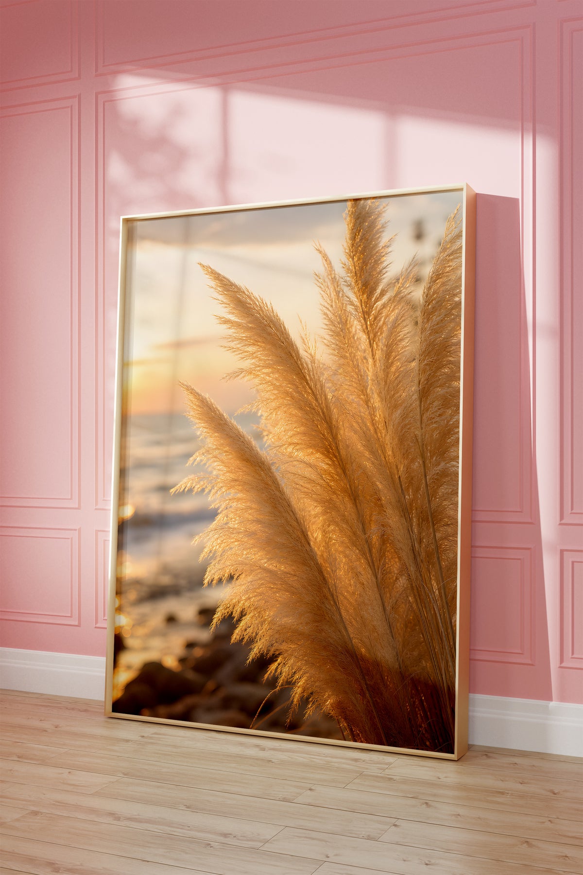 Motiv mit goldenen Pampaswedeln in Nahaufnahme am Strand im warmen Abendlicht, als gerahmtes Poster vor einer rosafarbenen Wand.