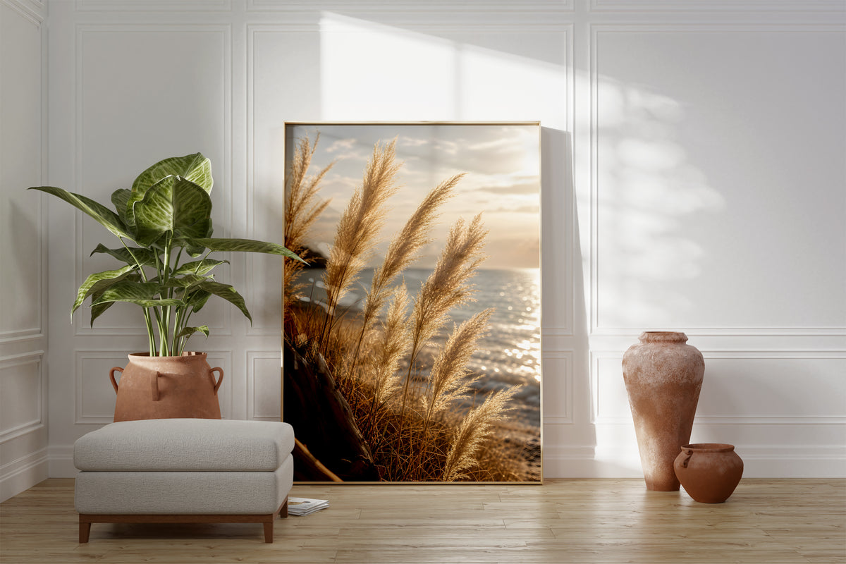 Motiv mit goldenen Pampasgräsern vor glitzerndem Meer im Abendlicht, als gerahmtes Poster neben Sessel und Zimmerpflanze.
