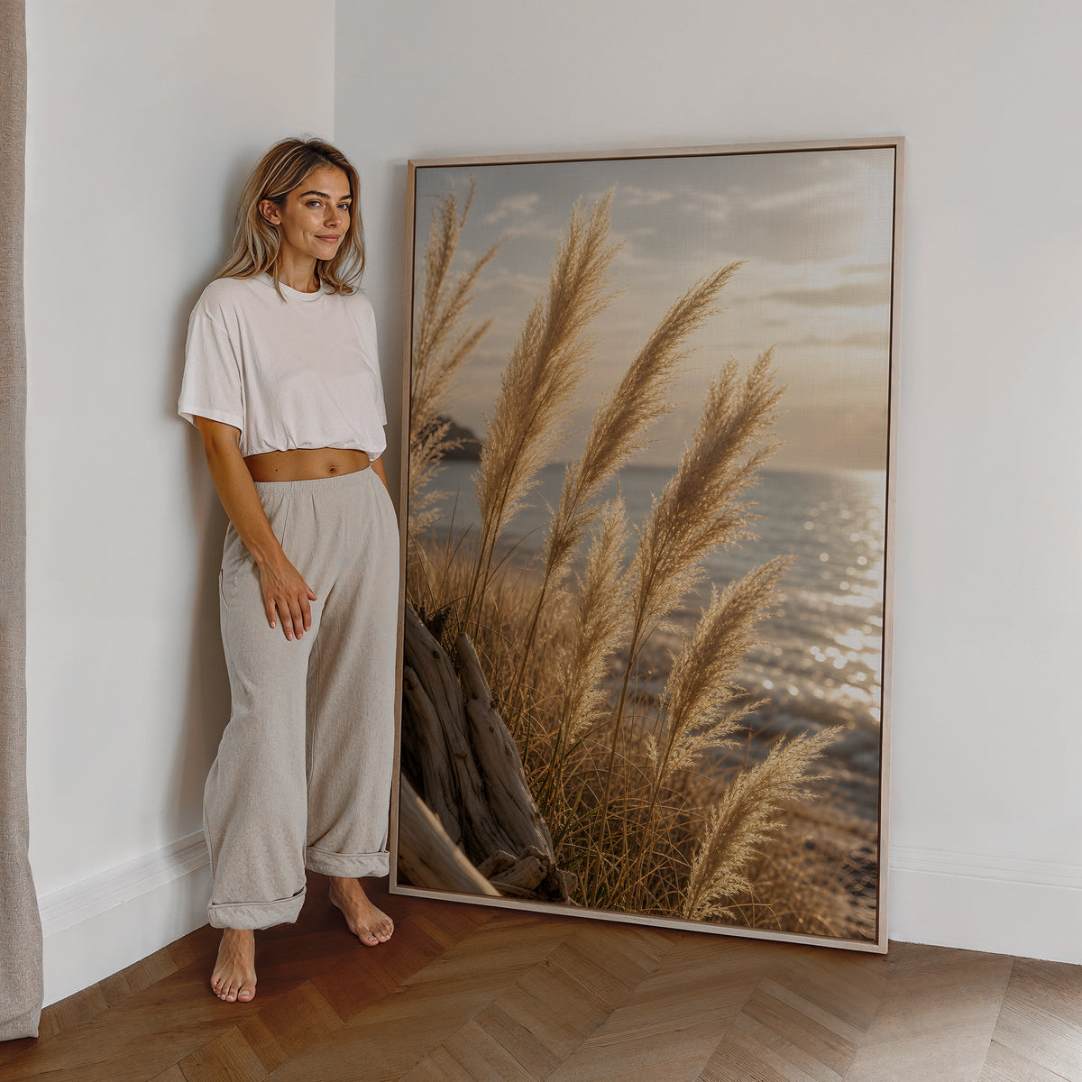 Motiv mit goldenen Pampasgräsern vor glitzerndem Meer im Abendlicht, als großes gerahmtes Wandbild neben einer Frau.