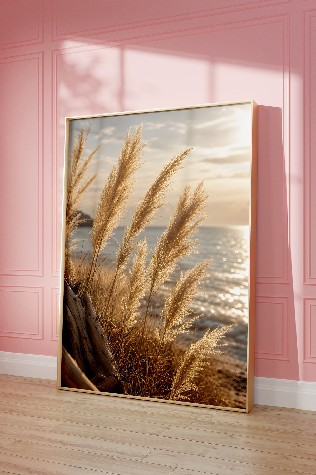 Motiv mit goldenen Pampasgräsern vor glitzerndem Meer im Abendlicht, als gerahmtes Poster vor einer rosafarbenen Wand.