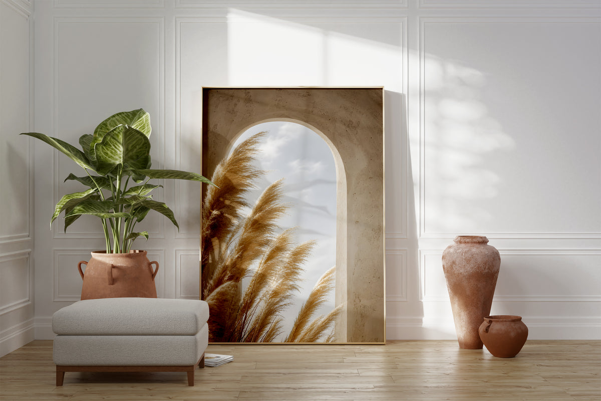 Motiv mit goldenen Pampaswedeln vor einem sandfarbenen Rundbogen mit Blick zum Himmel, als gerahmtes Poster neben Sessel und Zimmerpflanze.