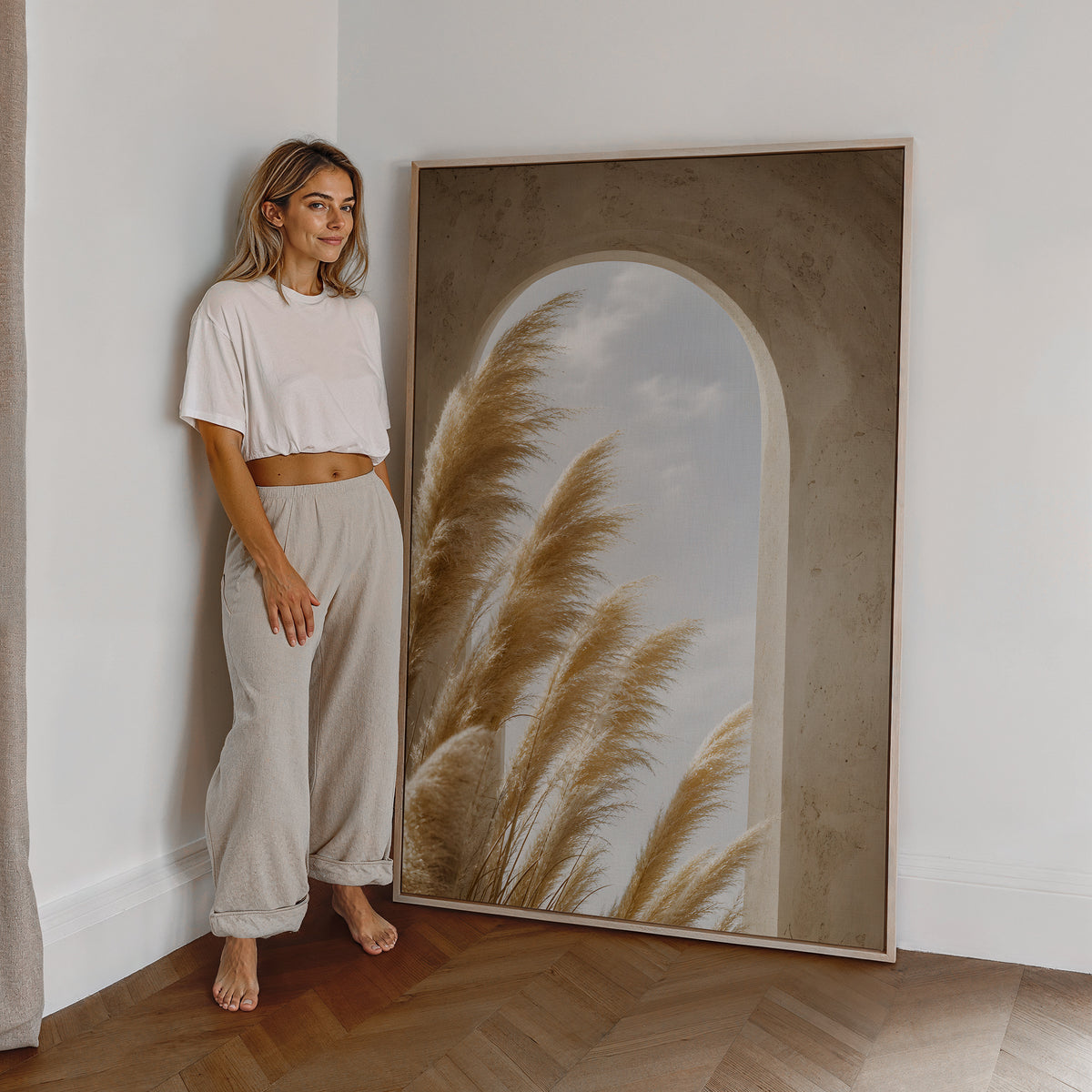 Motiv mit goldenen Pampaswedeln vor einem sandfarbenen Rundbogen mit Blick zum Himmel, als großes gerahmtes Wandbild neben einer Frau.