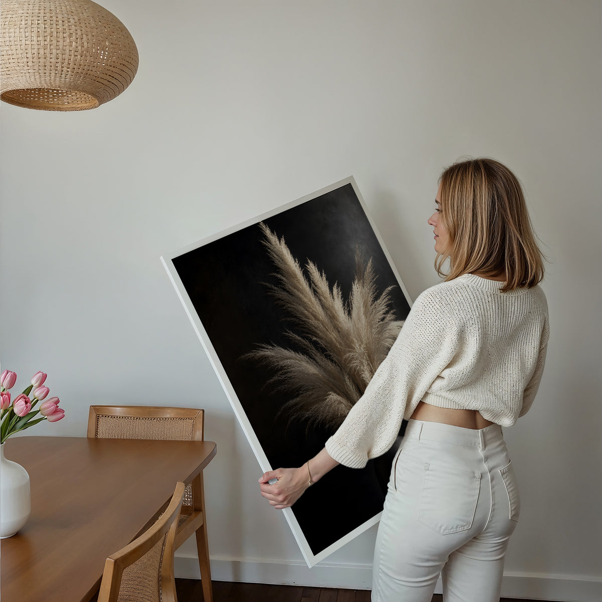 Motiv mit einem hellen Pampasbouquet in einer schwarzen Vase vor schwarzem Hintergrund, über einem Bett im hellen Schlafzimmer.