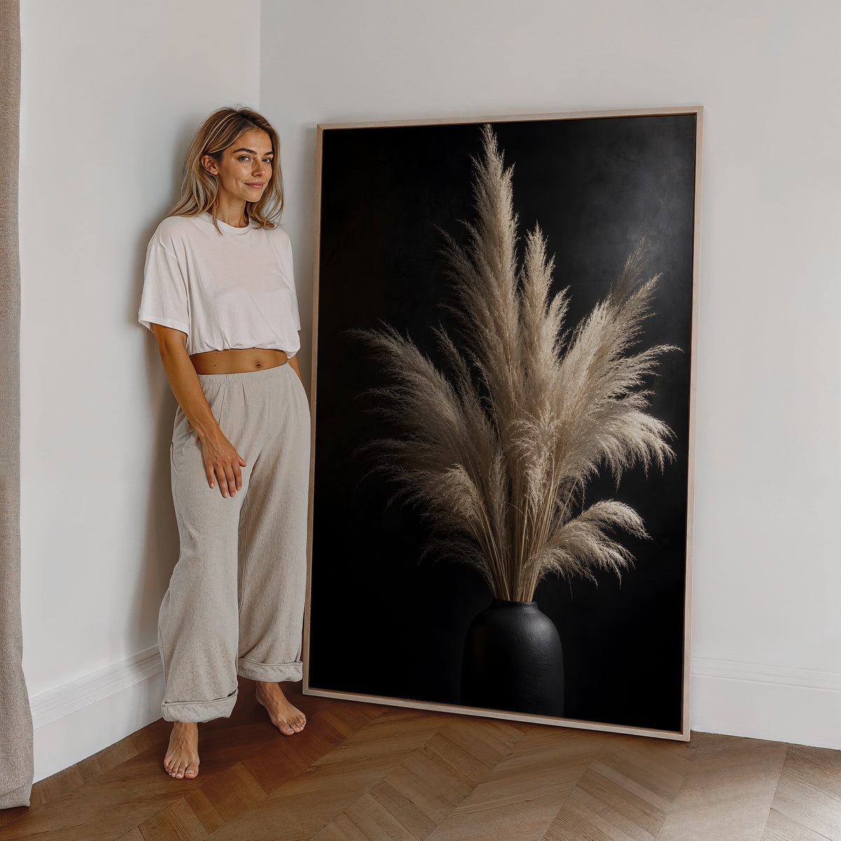 Motiv mit einem hellen Pampasbouquet in einer schwarzen Vase vor schwarzem Hintergrund, als großes gerahmtes Wandbild neben einer Frau.