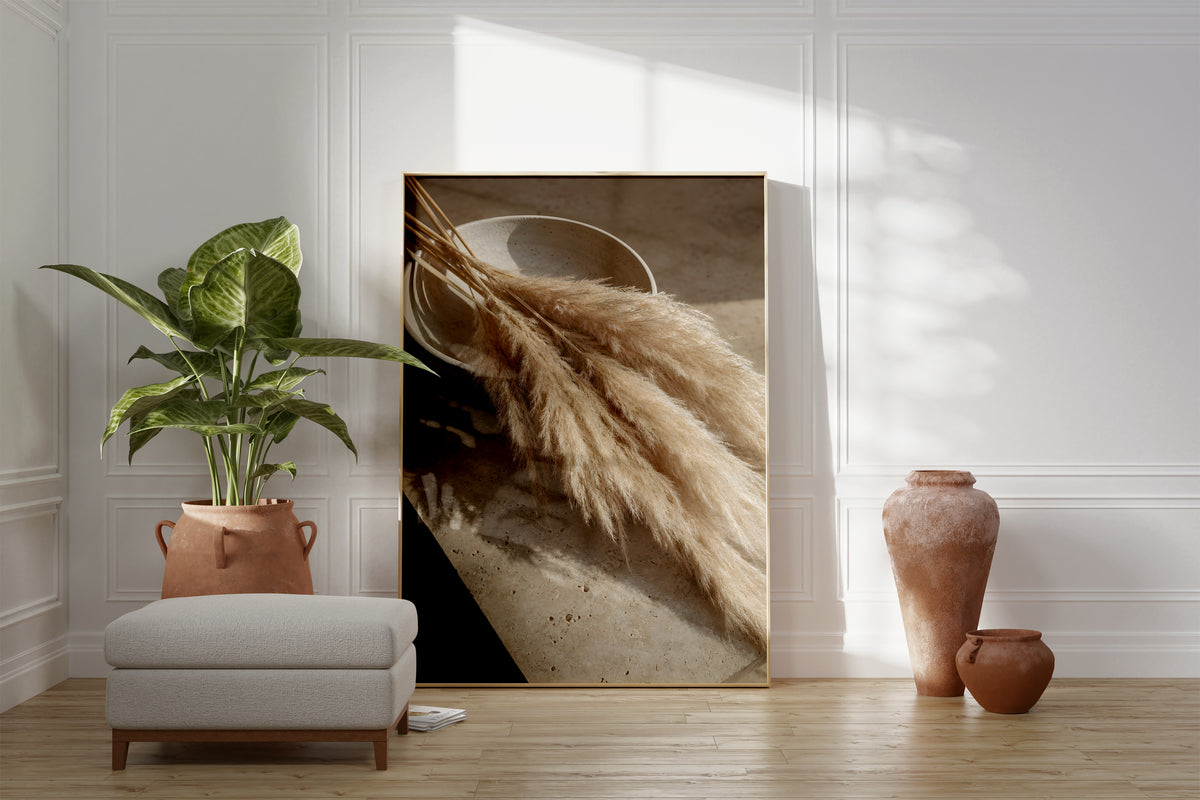 Motiv mit einem liegenden Pampasbouquet auf einer Steinfläche neben einer Keramikschale, als gerahmtes Poster neben Sessel und Zimmerpflanze.