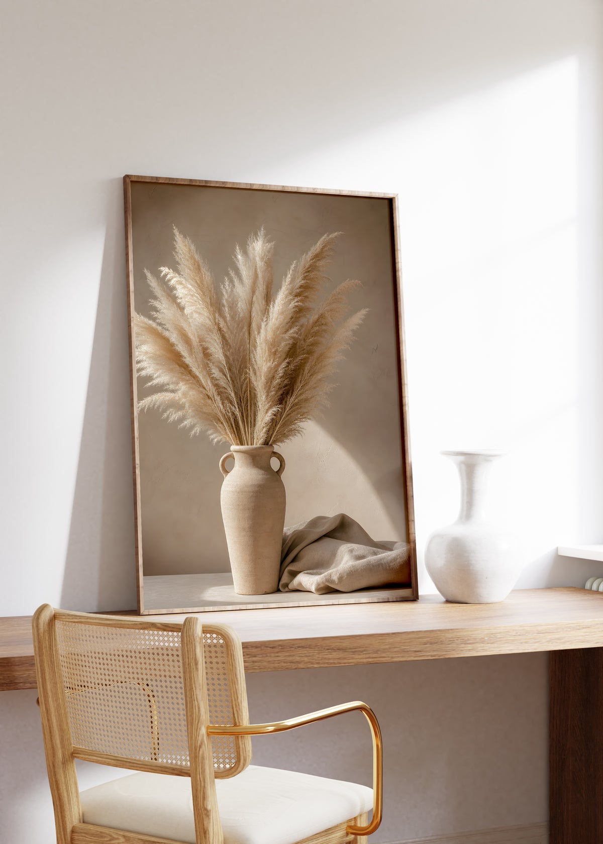 Gerahmtes Poster mit einem Pampasbouquet in einer beigefarbenen Henkelvase neben hellem Stoff, auf einem Holztisch neben einer weißen Vase.