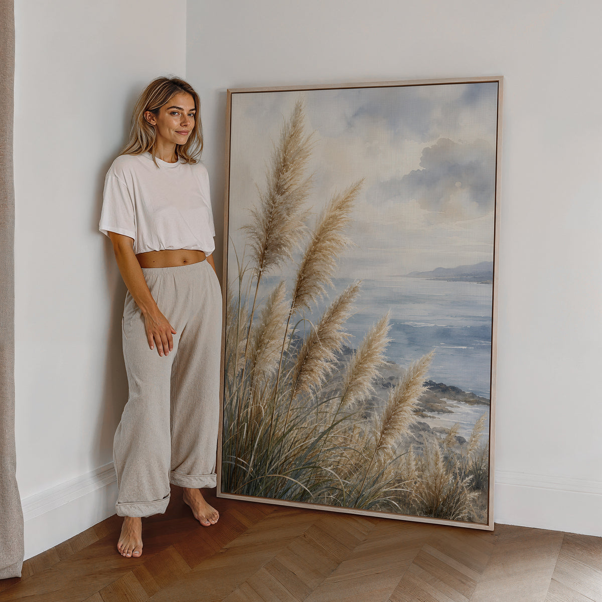 Motiv mit Pampasgräsern an einer blauen Küste unter Wolken in Aquarelloptik, als großes gerahmtes Wandbild neben einer Frau.