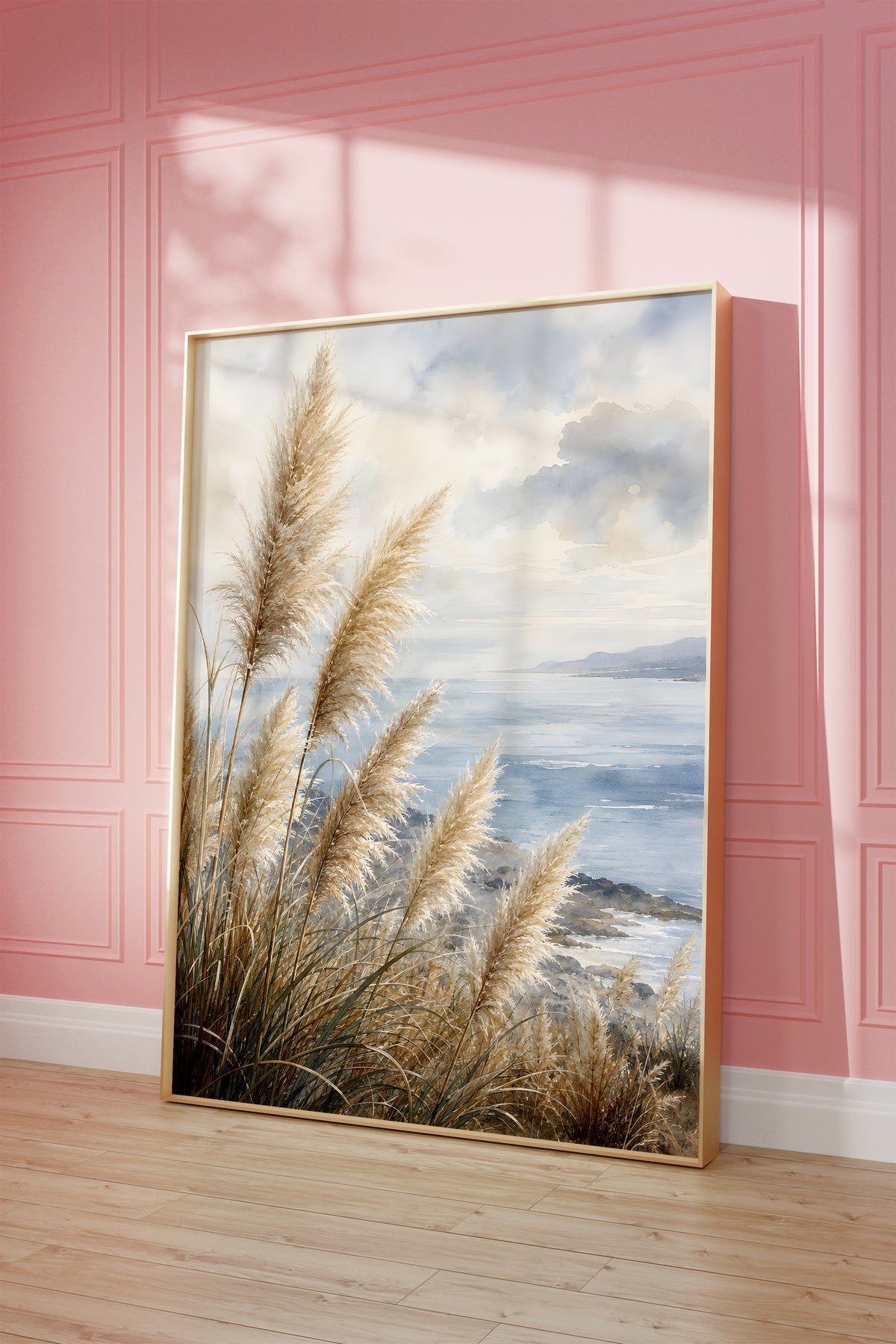 Motiv mit Pampasgräsern an einer blauen Küste unter Wolken in Aquarelloptik, als gerahmtes Poster vor einer rosafarbenen Wand.