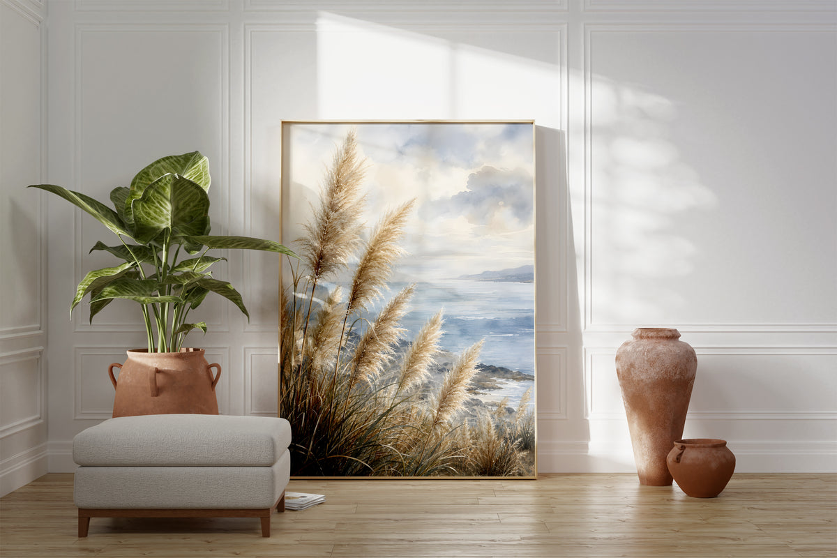 Motiv mit Pampasgräsern an einer blauen Küste unter Wolken in Aquarelloptik, als gerahmtes Poster neben Sessel und Zimmerpflanze.