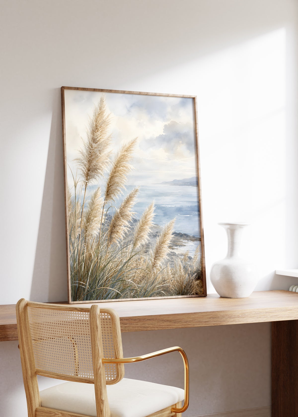 Gerahmtes Poster mit Pampasgräsern an einer blauen Küste unter Wolken in Aquarelloptik, auf einem Holztisch neben einer weißen Vase.