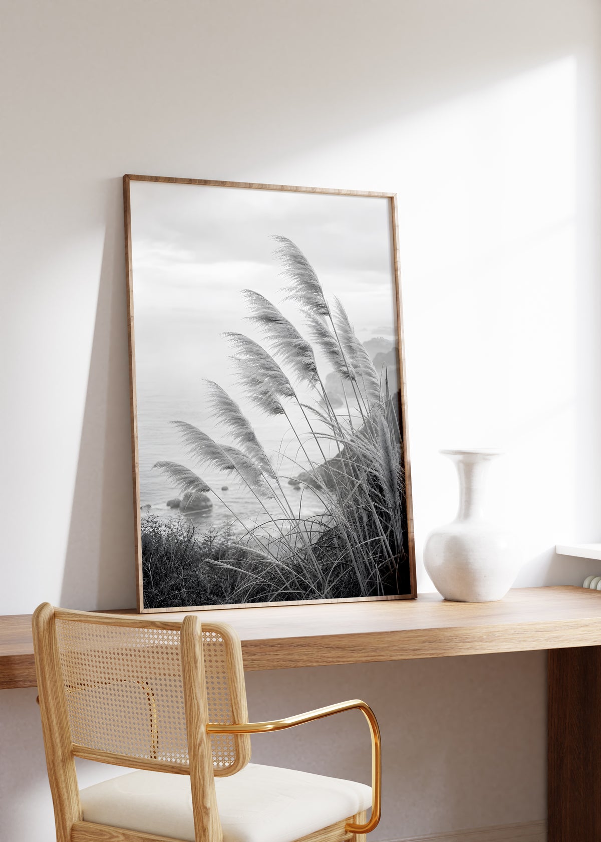 Gerahmtes Poster mit Pampasgräsern an einer Küste in Schwarz-Weiß, auf einem Holztisch neben einer weißen Vase.