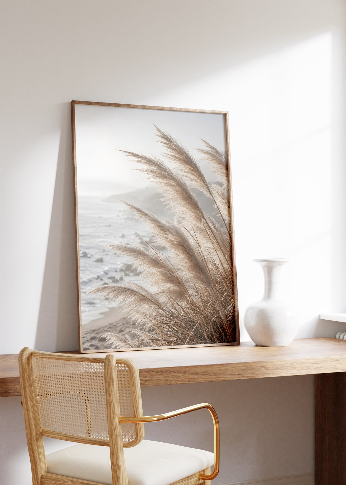 Gerahmtes Poster mit hellen Pampasgräsern vor einer nebligen felsigen Küste, auf einem Holztisch neben einer weißen Vase.