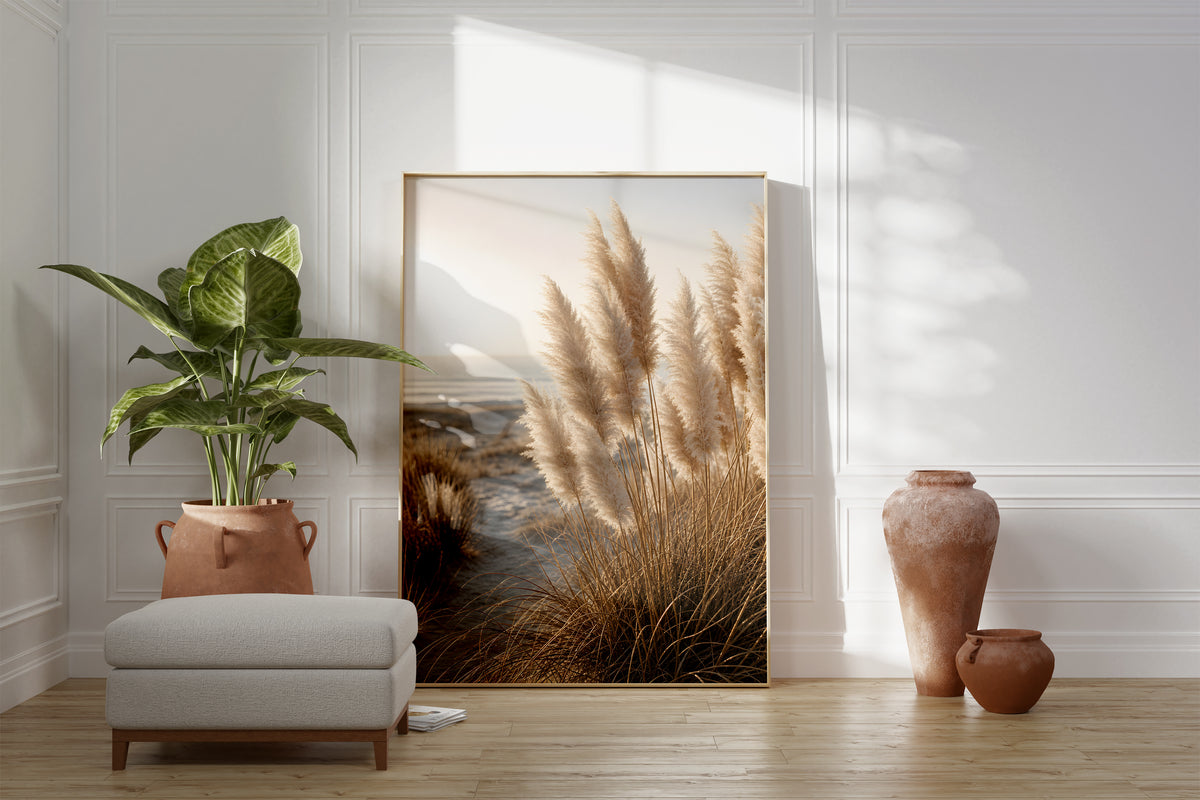 Motiv mit hellen Pampasgräsern in sandigen Dünen am Meer, als gerahmtes Poster neben Sessel und Zimmerpflanze.