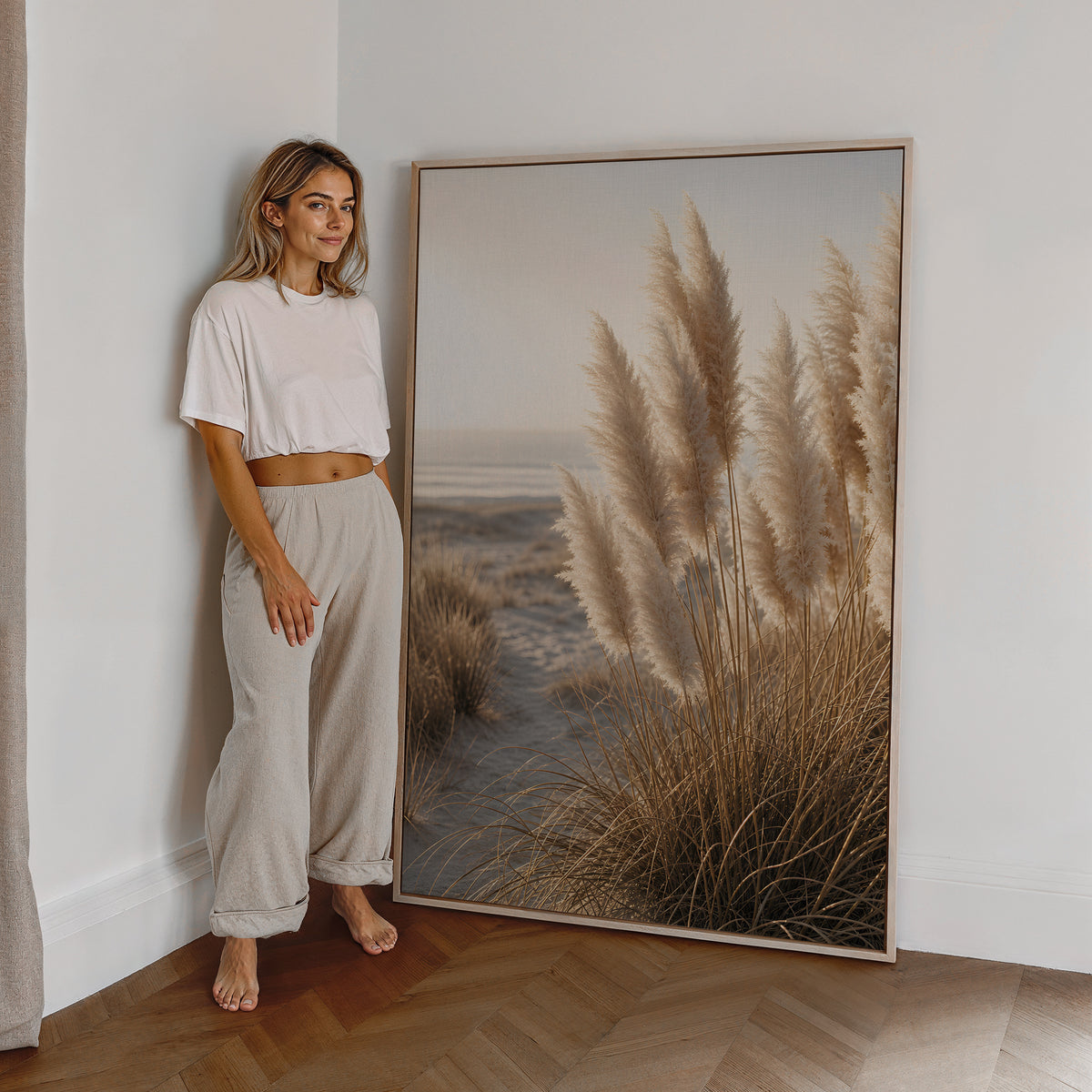 Motiv mit hellen Pampasgräsern in sandigen Dünen am Meer, als großes gerahmtes Wandbild neben einer Frau.