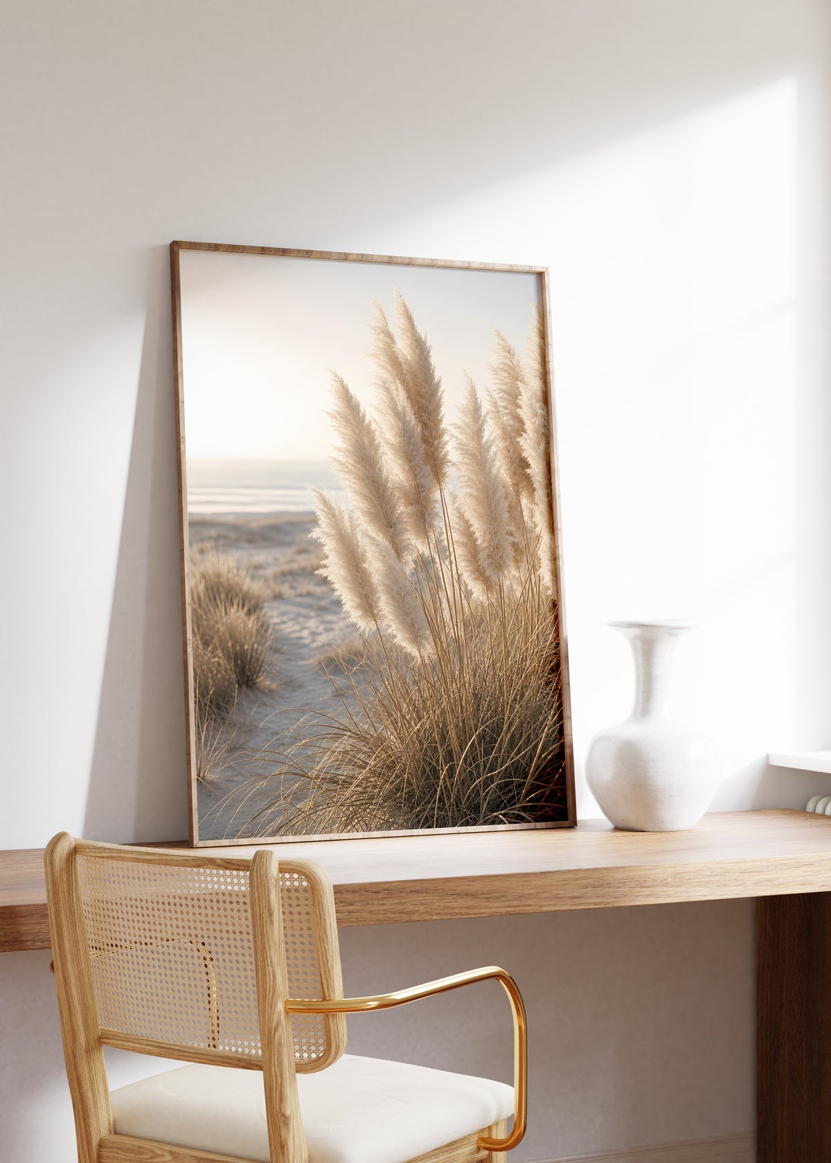 Gerahmtes Poster mit hellen Pampasgräsern in sandigen Dünen am Meer, auf einem Holztisch neben einer weißen Vase.