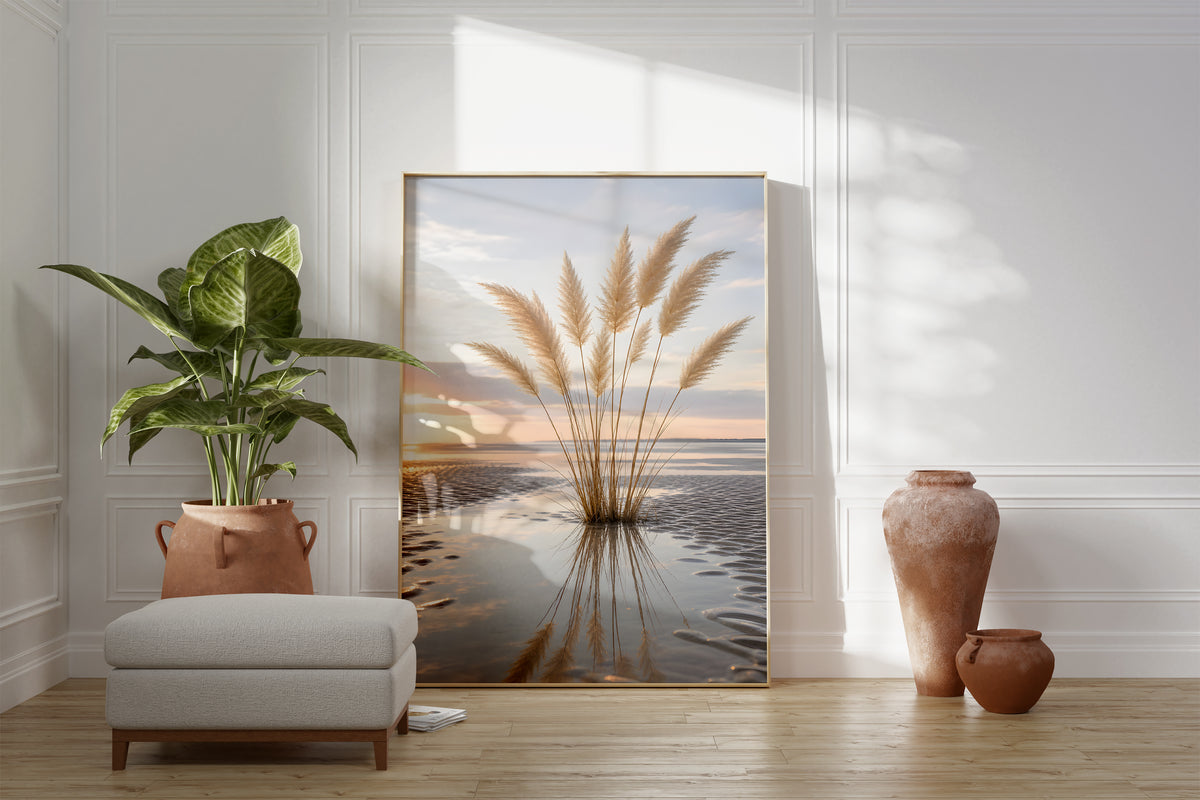 Motiv mit einem Pampasgrasbüschel mit Spiegelung im nassen Strandsand bei Sonnenuntergang, als gerahmtes Poster neben Sessel und Zimmerpflanze.