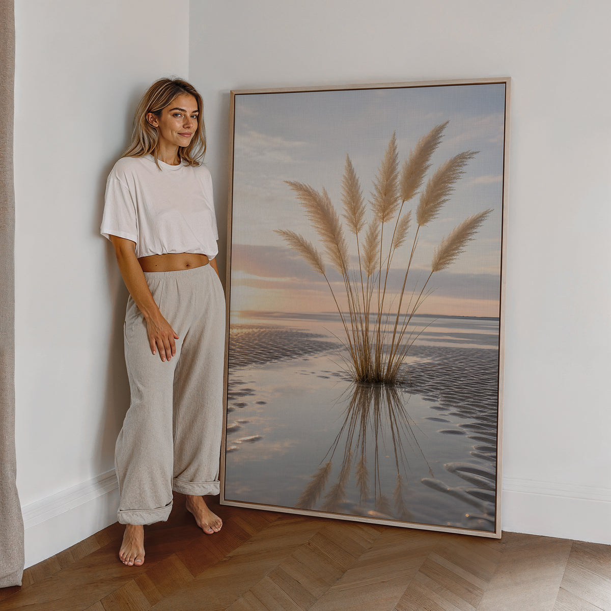 Motiv mit einem Pampasgrasbüschel mit Spiegelung im nassen Strandsand bei Sonnenuntergang, als großes gerahmtes Wandbild neben einer Frau.