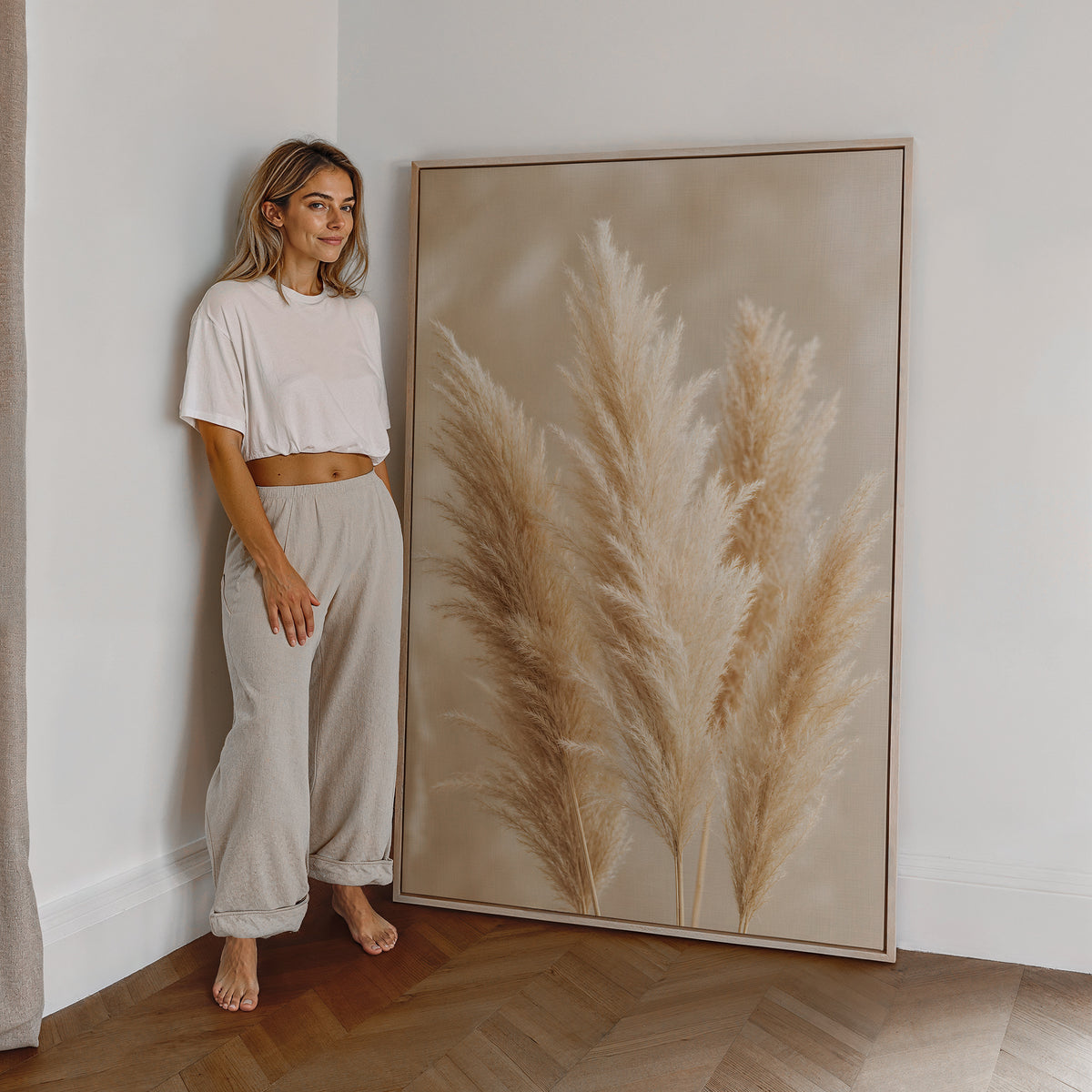 Motiv mit mehreren flauschigen Pampaswedeln in Creme und Beige vor weich verschwommenem Hintergrund, als großes gerahmtes Wandbild neben einer Frau.
